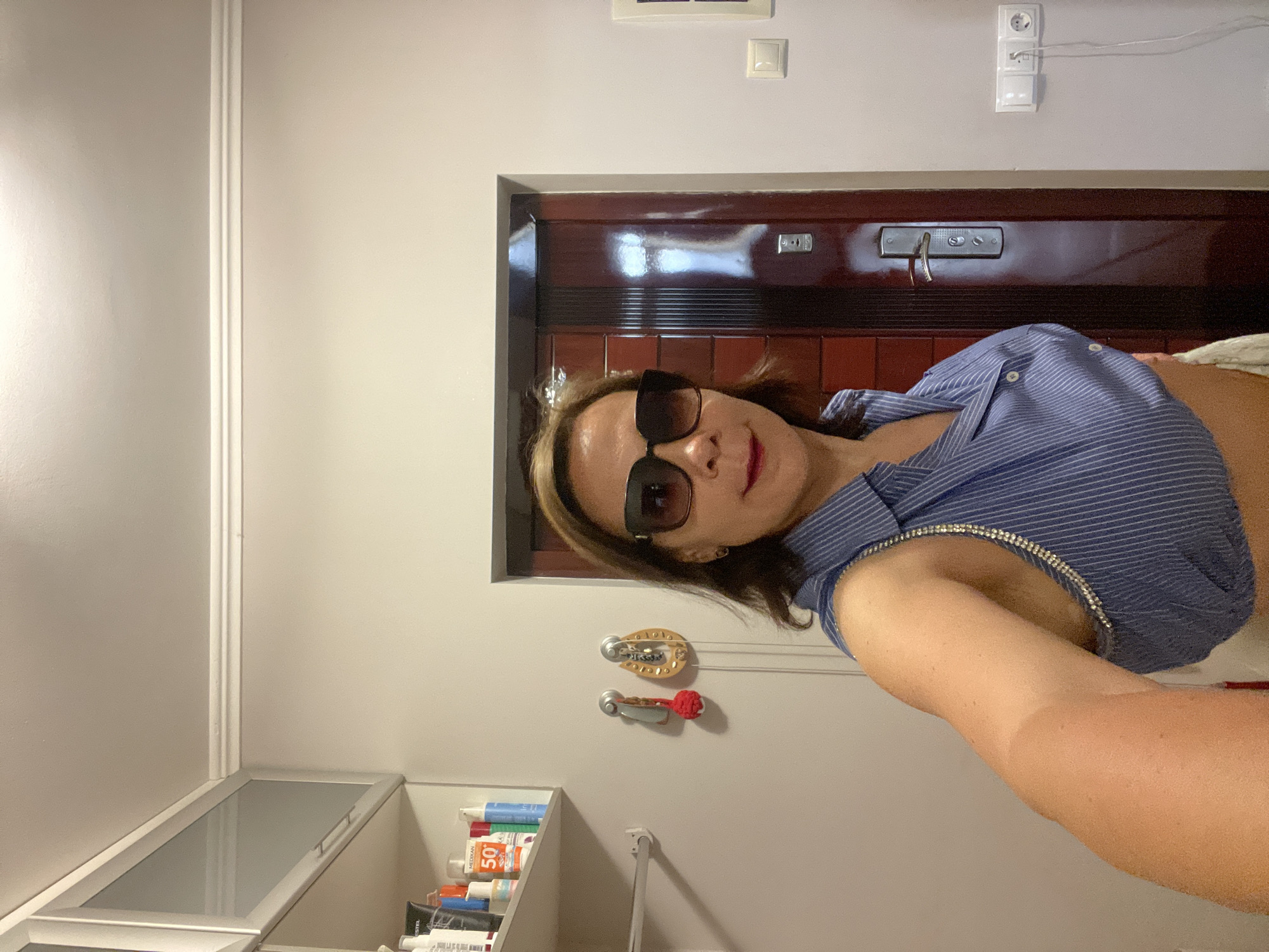 A medium-skinned woman with dark hair and sunglasses poses for a selfie indoors. She is wearing a blue pinstriped shirt with a sparkly trim along the neckline. Behind her, a dark wooden door with a silver handle is visible, as are shelves with various toiletries, including a sunscreen bottle labeled "50". The room appears to be a bathroom or dressing area, with white walls and molding near the ceiling. There are also electrical outlets visible on the wall. The overall setting is domestic and unremarkable.