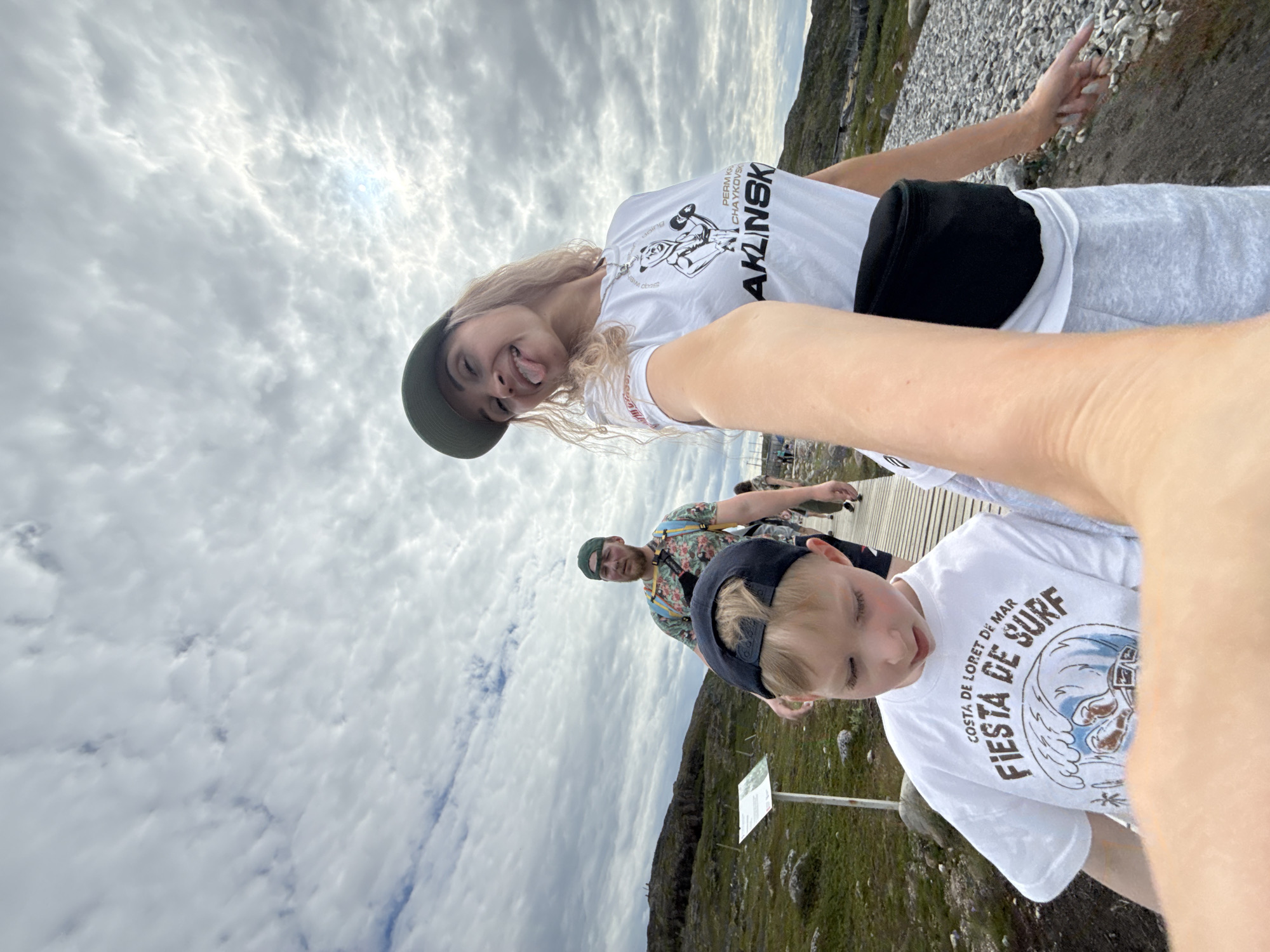 This image captures a group of people outdoors in Teriberka, Russia. The sky is overcast with textured clouds. In the foreground, a young boy wearing a white t-shirt with "Fiesta de Surf" printed on it and a black baseball cap is looking down. To his right, a woman wearing a white t-shirt with graphics, a green baseball cap, and a black fanny pack is sticking her tongue out at the camera, holding her arm out to take a selfie. Behind them, a man in a floral-patterned t-shirt and a green baseball cap walks along a wooden boardwalk. The environment appears to be a rocky, somewhat barren landscape with patches of green grass and low-lying vegetation. A white sign is visible on a post to the left of the boardwalk. The overall scene suggests a casual outdoor excursion in a coastal or subarctic region.