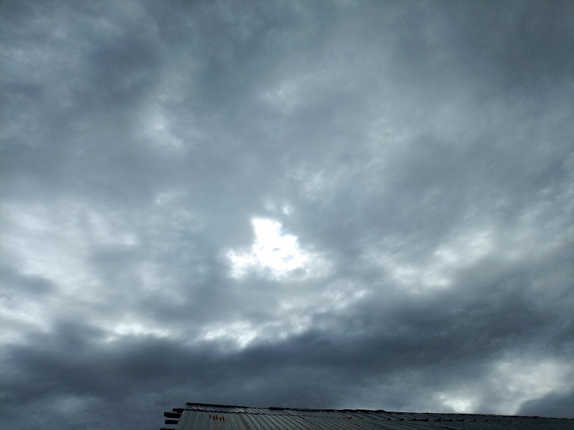 The image depicts a cloudy sky above a metal roof in the city of Kyzyl-Suu, Kyrgyzstan. The sky is predominantly overcast with shades of gray, but a bright patch of sunlight breaks through the clouds in the upper center of the frame. The roof, visible in the lower portion of the image, appears to be corrugated metal and shows signs of weathering, with some rust spots visible. The overall scene suggests a potentially gloomy or approaching rainy weather, with the sun attempting to penetrate the dense cloud cover. No people or animals are present in the image, and no specific actions or interactions are depicted. The environment is urban, indicated by the presence of a man-made structure (the roof).