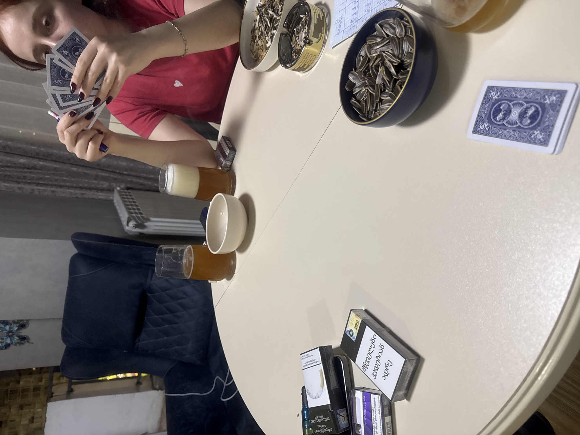 Here's a forensic description of the image:
**Scene:** A round, light-colored table is the central focus. An individual is seated at the table, holding a hand of playing cards with blue backs. Several items are arranged on the table: bowls containing sunflower seeds (some shelled, some unshelled), glasses/containers of liquid (possibly beer or juice), cigarette packs (some displaying text in Georgian), and loose playing cards. A dark blue, tufted chair is visible on the left side of the table.
**People:** One person is partially visible in the frame, holding playing cards. Details like skin tone, nail polish color (dark purple), and the color of the shirt (red) are discernible.
**Objects:**
* Playing cards (blue backs).
* Bowls: containing sunflower seeds, some with shells and some without
* Beverage glasses/containers: Filled with a liquid, possibly beer.
* Cigarette packs: Several brands are visible, some with Georgian script.
* White Ceramic Bowl
* A dark blue, tufted chair.
**Actions/Interactions:** The person is actively holding and presumably looking at the playing cards.
**Environment:** The background is somewhat blurred, but includes a window and possibly a curtain. The scene appears to be indoors, possibly a home.
**Location:** Based on the Georgian text on the cigarette packs, it's likely that the location is in Tbilisi, Georgia.