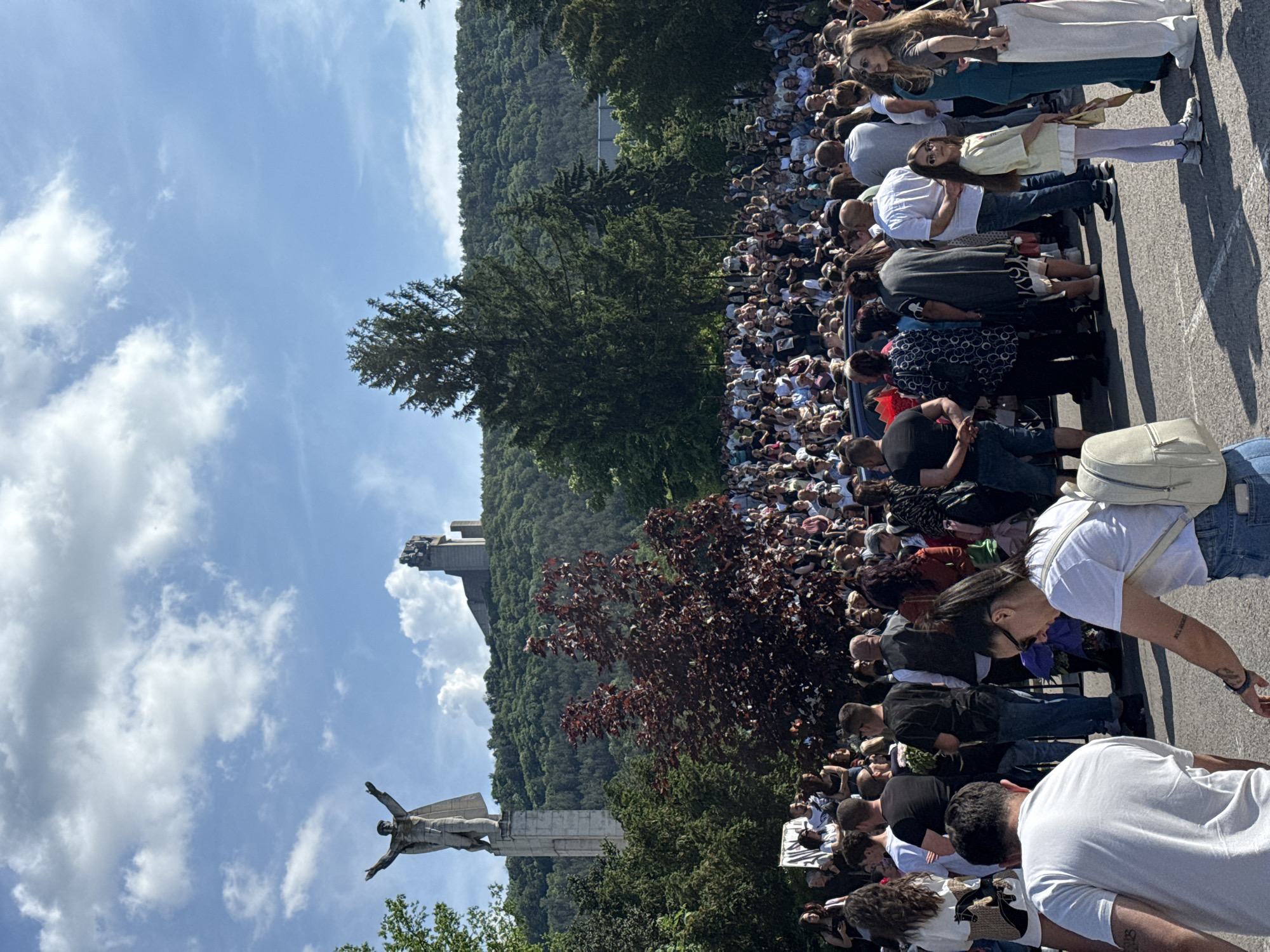 The image shows a large crowd of people gathered in Shumen, Bulgaria, near the Monument to 1300 Years of Bulgaria. The monument features a towering statue of a figure with outstretched arms. The environment includes a hillside covered in trees and a partially cloudy sky. People are standing or sitting, looking toward the monument.