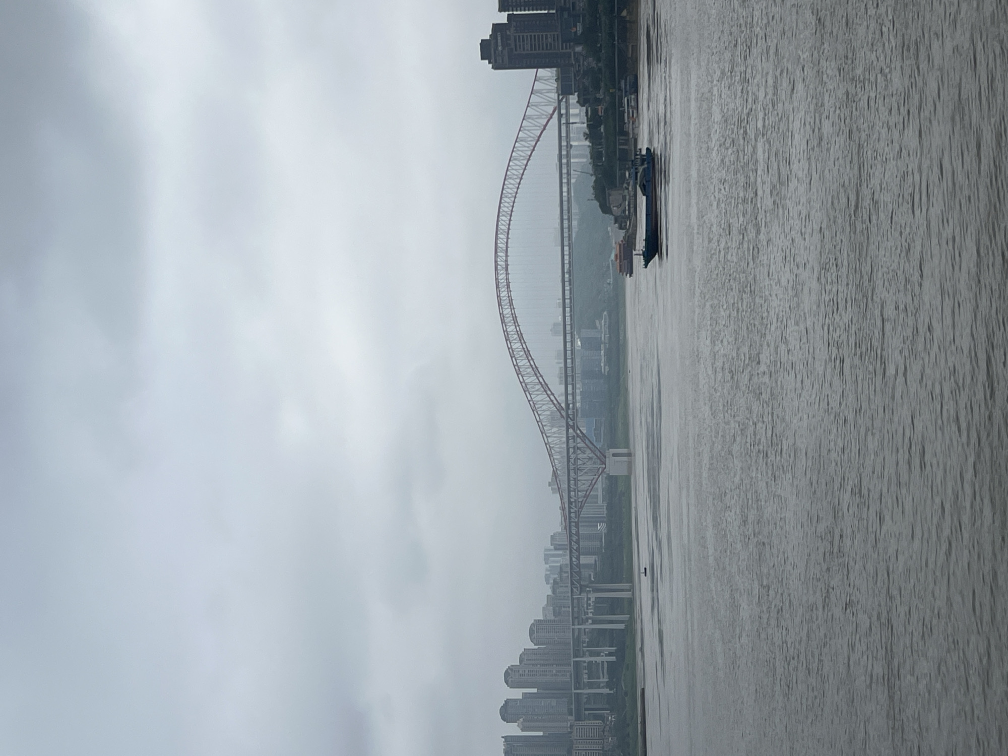 A hazy, overcast sky hangs above a wide river in Yuzhong District, China. The river's surface is choppy, reflecting the muted tones of the sky. Several boats are visible on the water, including a long, blue barge in the foreground and other vessels further in the distance. On the left bank, a cluster of tall, modern buildings rises, forming a cityscape. A prominent bridge with a distinctive arch spans the river, connecting the banks. Another, more utilitarian bridge with numerous supporting pillars is visible behind it. The architecture suggests a dense urban environment.