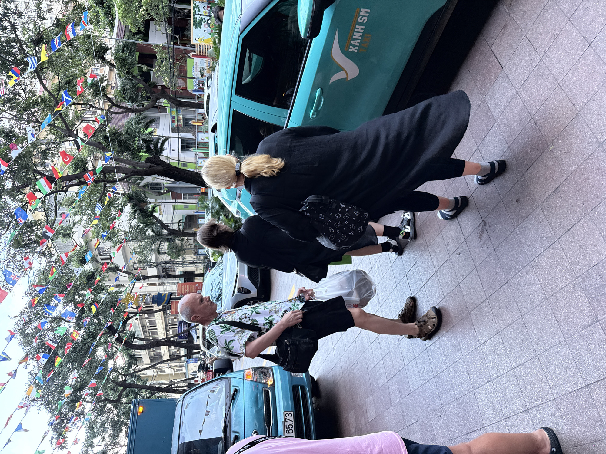 In Nha Trang Ward, Vietnam, a man in a white floral shirt and black shorts stands on a sidewalk, holding a black shoulder bag and a clear plastic bag containing items. He is facing away from the camera, looking towards the street. To his left, a teal-colored truck with the license plate "65.73" is parked. To his right, a teal-colored taxi with "XANH SM TAXI" written on the side is visible. Behind the man, two women are walking, one in a black outfit with a black bag and the other in a black dress. Overhead, numerous small flags from various countries are strung between trees. Buildings and more trees are visible in the background, suggesting an urban setting. The ground is paved with square tiles.