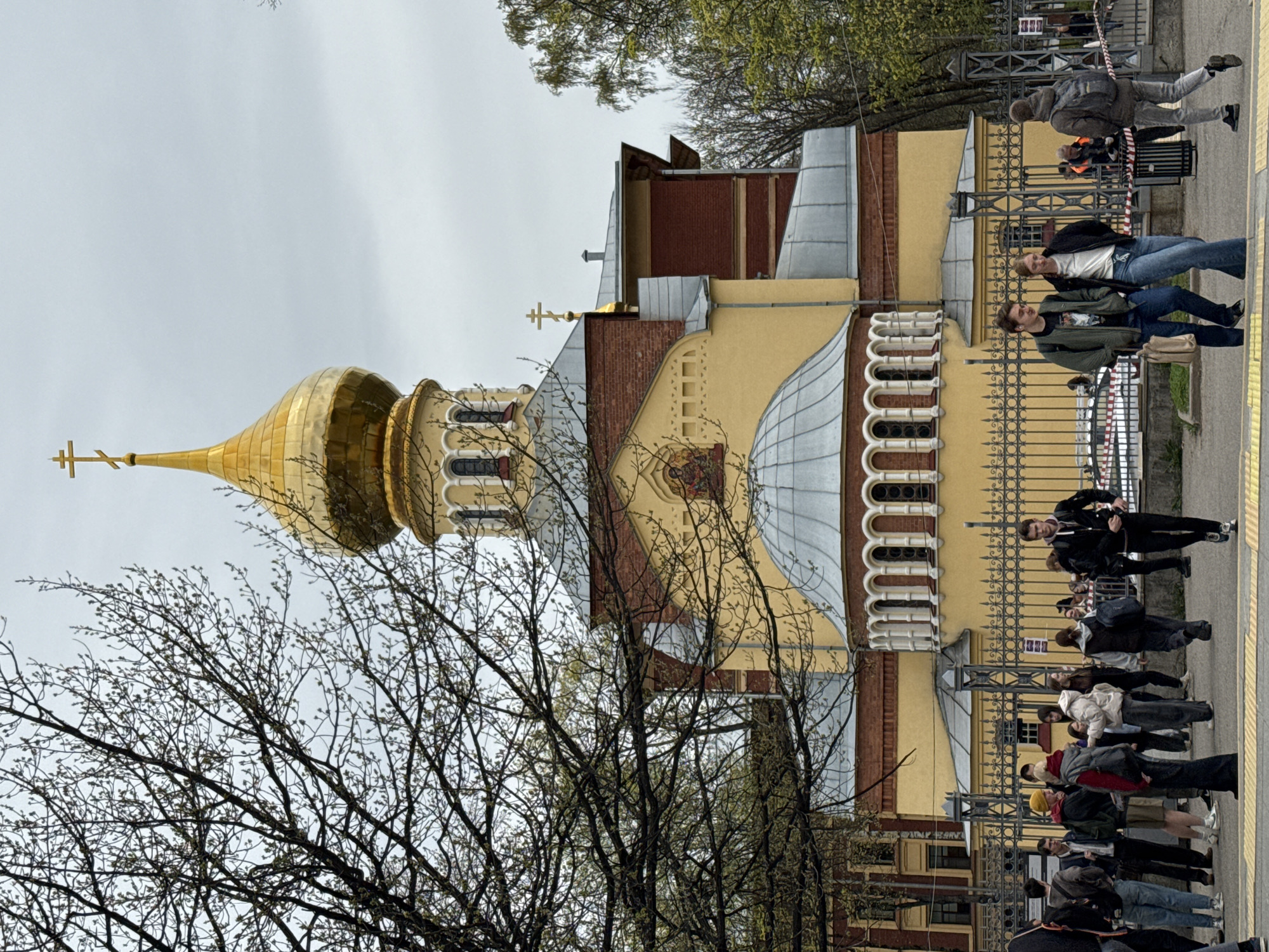 The image shows a partial view of the Saint John Chrysostom Church in Saint Petersburg, Russia. The church has a golden dome topped with a cross. A leafless tree occupies the foreground. A line of people walks along a sidewalk next to a metal fence that runs along the side of the church. The sky is overcast.