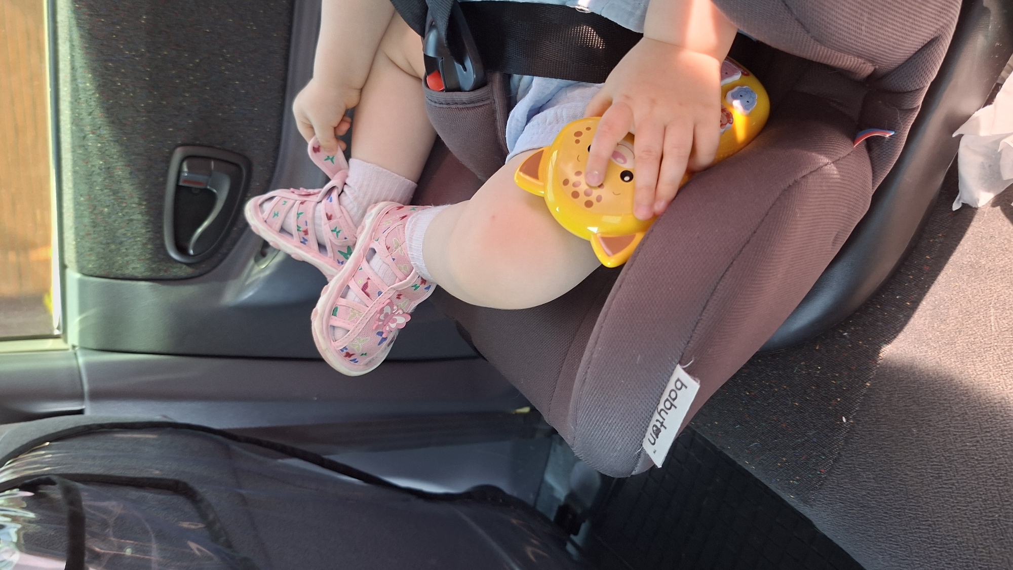 A child sits buckled in a gray car seat, holding a yellow giraffe-shaped toy. The child wears light pink shoes with floral embroidery and light pink socks. The car seat is placed inside a car, with the door panel and handle visible on the left. A tag reading "babyton" is attached to the car seat. The location is Прикамский, Russia, based on provided information.