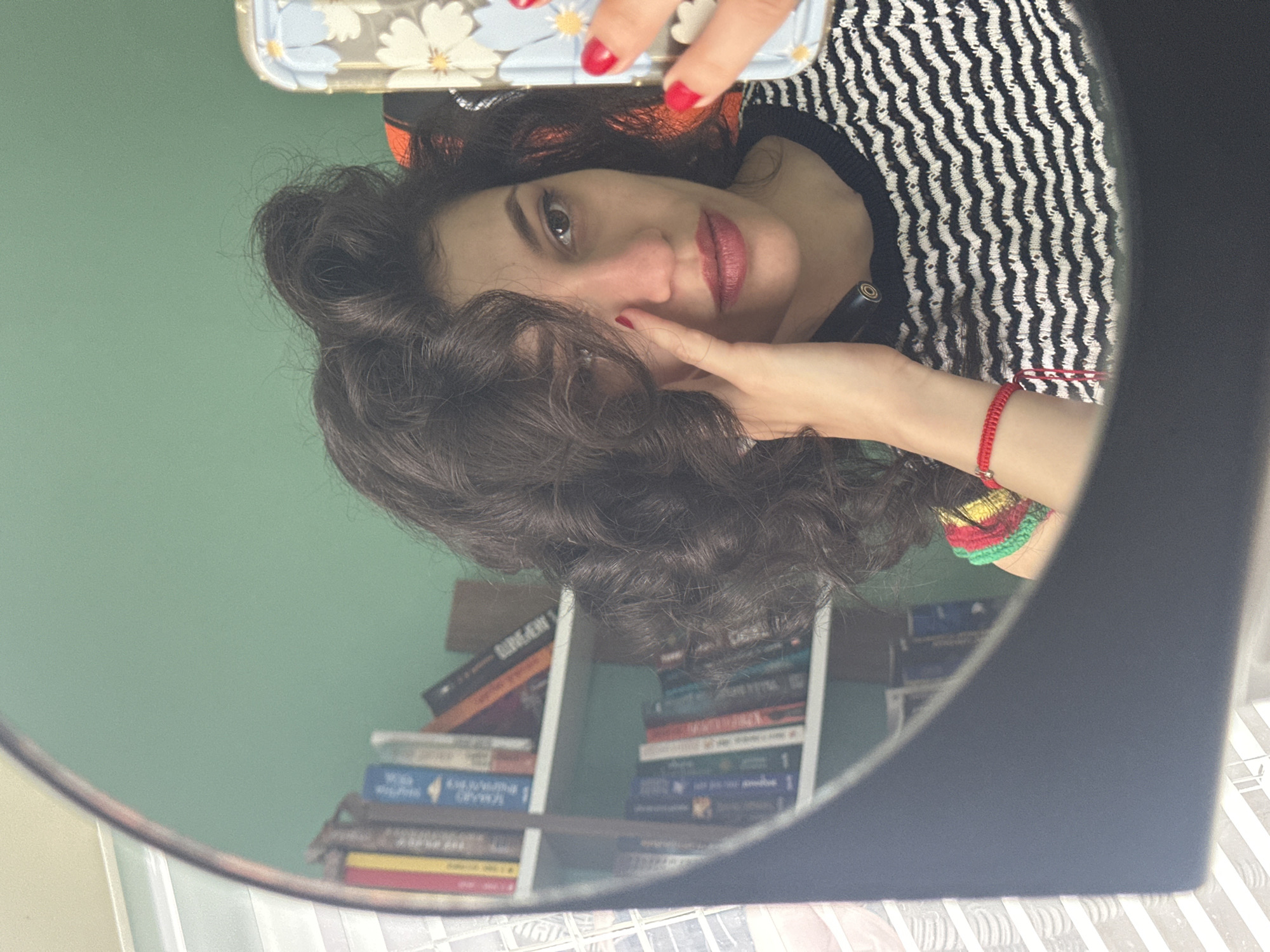 A young woman with curly dark hair is taking a selfie in a mirror. She is wearing a black and white striped shirt with a crocheted neckline. Her left hand is touching her face, and she is wearing a red bracelet. In the reflection of the mirror, bookshelves filled with books are visible, along with a glimpse of what appears to be a white tiled wall. The background of the room is painted a light green. The image does not provide any information about the location being in Sofia, Bulgaria.