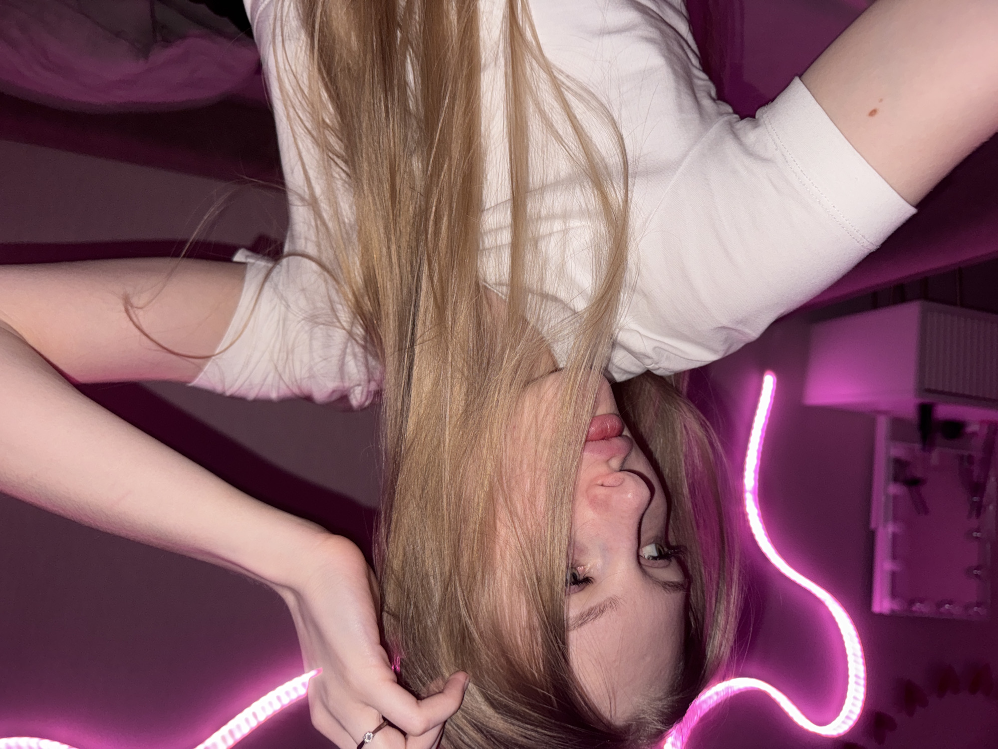 A young woman with long blonde hair and light-colored eyes is pictured from the chest up, looking to her left. She is wearing a white t-shirt. Her right hand is raised to her head, touching her hair. A pink neon light, shaped like a heart, is visible behind her. The background appears to be a room with a dresser or vanity to the left and a purple couch or bed behind her. The image was taken in a bedroom at night. There is no indication that this image was taken in the city of Perm, Russia.