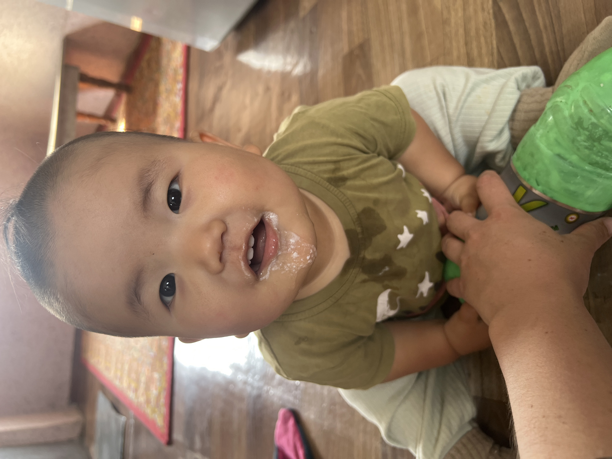 The image shows a close-up, upside-down view of a baby. The baby has a messy mouth with white residue around it. The baby is wearing a green t-shirt with white stars. The background appears to be a wooden floor and some colorful patterned fabric. A person's hand is visible, holding a green object, possibly a bottle or toy. The overall impression is of a candid moment with a baby, likely during or after a meal. The lighting suggests it is daytime. The location is identified as Kara-Dıykan, Kyrgyzstan. There is no visible text in the image.