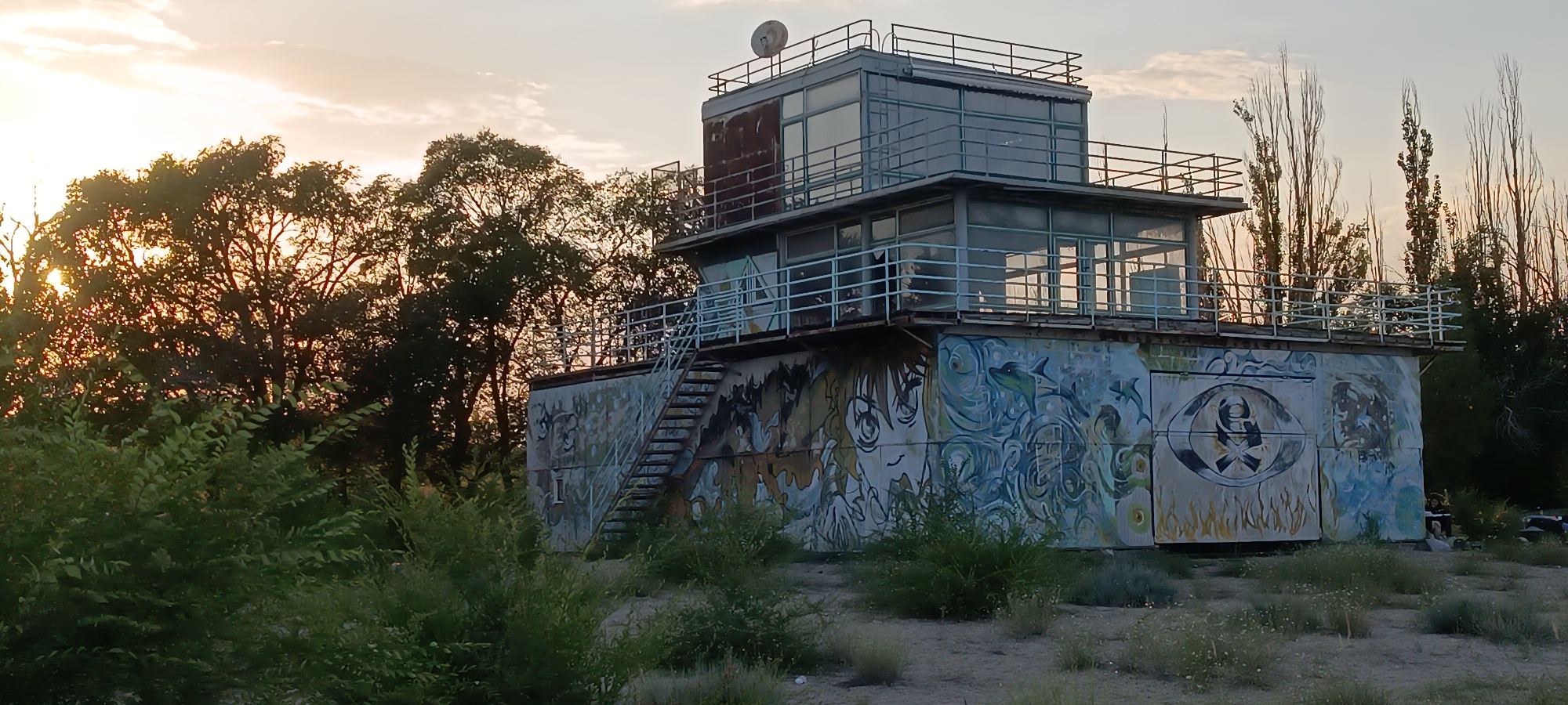 An abandoned, multi-story building, possibly a former control tower or observation post, stands amidst dry vegetation and tall, slender trees. The building's exterior is covered in colorful graffiti, including a large stylized eye with a skull and crossbones motif and depictions of anime characters and marine life. A metal staircase provides access to the upper levels, which feature large glass windows and metal railings. The sun is setting in the background, casting a warm glow on the scene and silhouetting some of the trees. The environment appears arid and somewhat neglected, suggesting a remote or undeveloped location. The specific location in Koshkol', Kyrgyzstan, is noted. There are no people visible in the image.