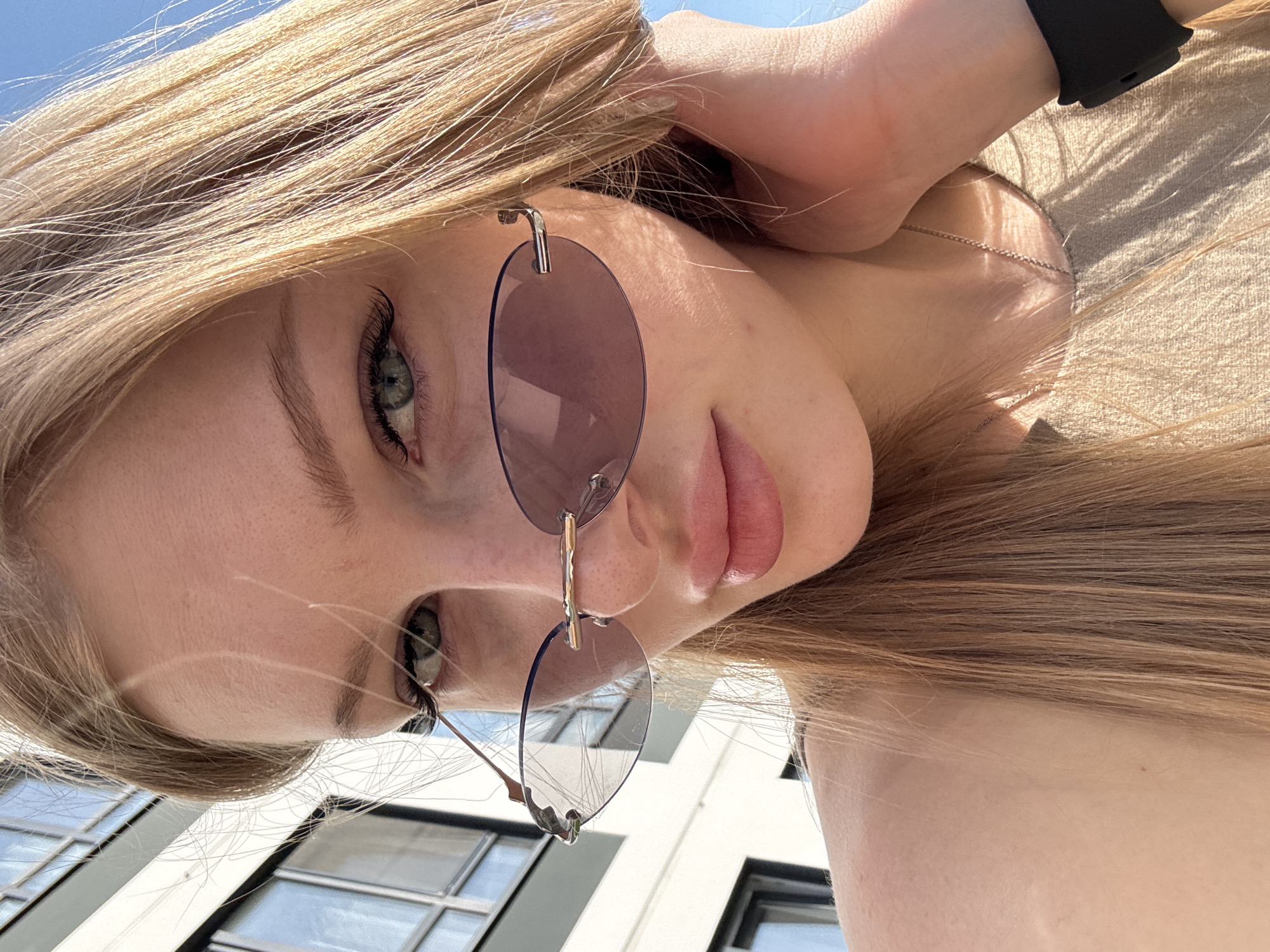 A young woman with long, blonde hair is photographed in a selfie-style shot. She is wearing rimless sunglasses with light purple lenses. The background appears to be a modern building with windows, suggesting an urban environment. Based on the buildings in the background the location might be in Perm, Russia. She has green eyes and is wearing a black smartwatch on her wrist. Her right hand is placed near the back of her head.