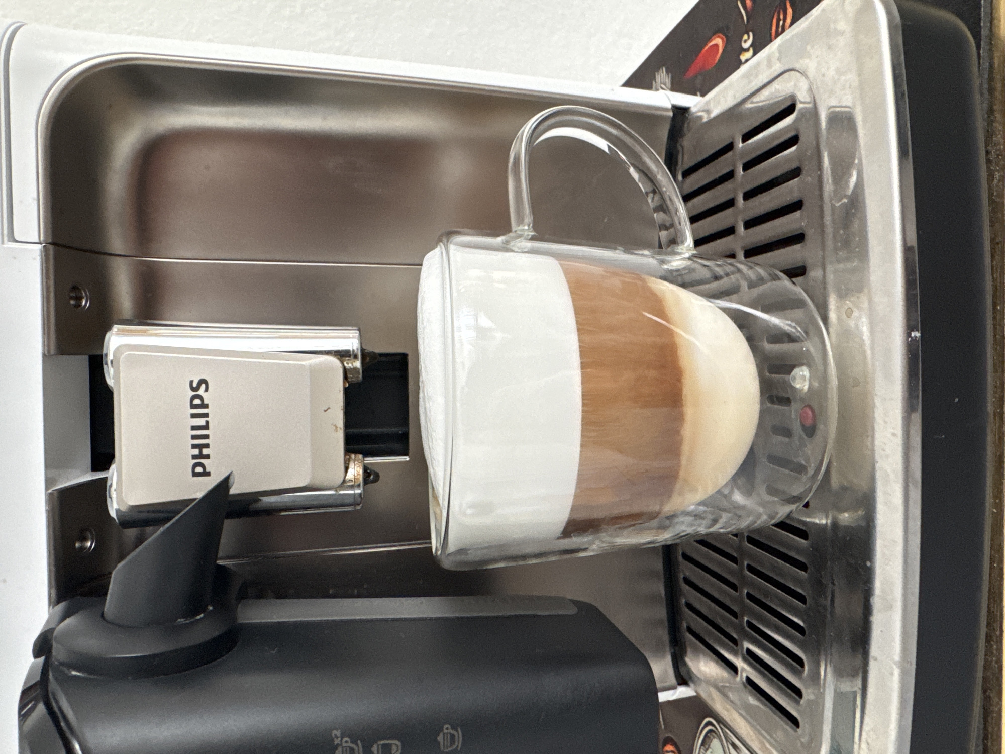 A clear glass mug filled with a layered coffee drink, topped with foamy milk, sits on the drip tray of a white and silver Philips coffee machine. The coffee machine is positioned in a domestic kitchen setting, likely in Rheine, Germany, as indicated by external knowledge about the user. The mug displays distinct layers of dark coffee at the bottom, a lighter brown coffee layer in the middle, and a thick white foam on top. The coffee machine has a black dispensing spout on the left and a silver panel with the Philips logo above it. The drip tray is made of brushed metal with parallel slats. The background shows a plain white wall.