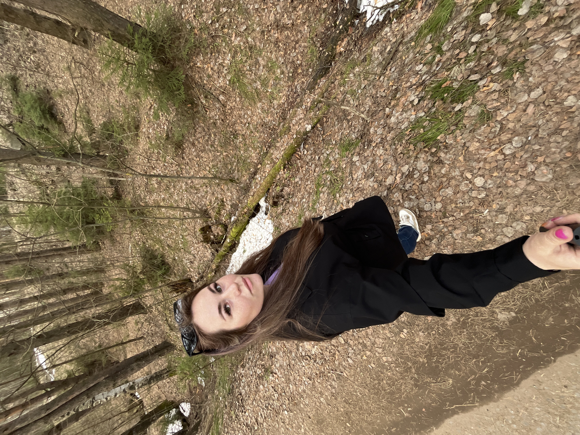 A young woman stands in a forest area in the city of Chaikovsky, Russia, during what appears to be early spring. The ground is covered with fallen leaves, with patches of grass and lingering snow present. She is wearing a dark jacket, blue jeans, and white sneakers. She has long brown hair and sunglasses perched atop her head. She is holding a mobile device, presumably taking a selfie. The environment consists of tall, bare trees and a path of some kind.