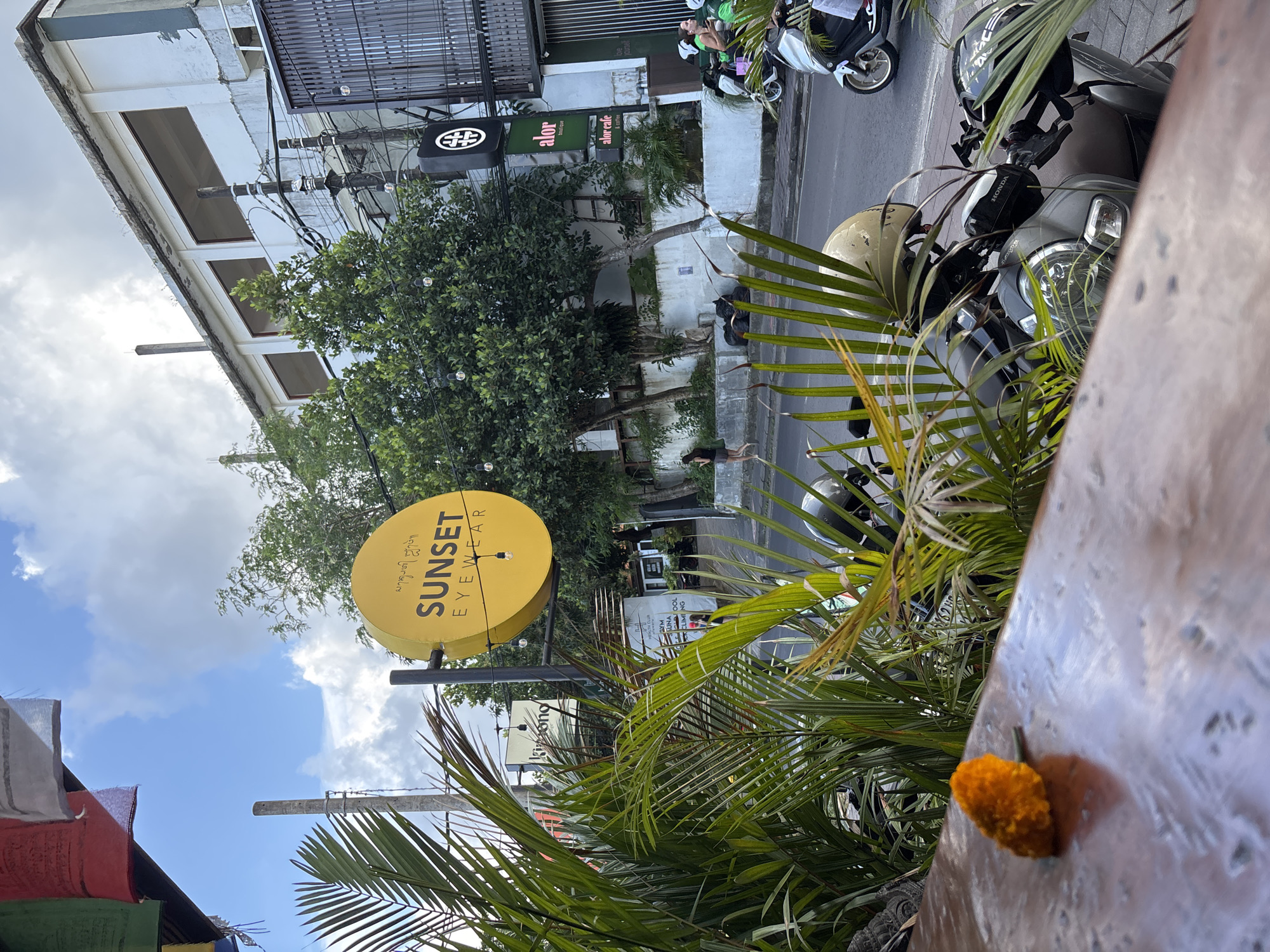 The image shows a street scene in Kuta Selatan, Indonesia. A yellow circular sign reading "SUNSET EYEWEAR" is visible in the foreground. Behind it, there are trees and a building, partially obscured. Parked scooters can be seen along the side of the road. In the foreground, palm leaves and an orange flower are visible. A sign that reads "alor" can be seen in the background. A section of a building is seen with windows and electrical wiring. The sky is visible with scattered clouds.