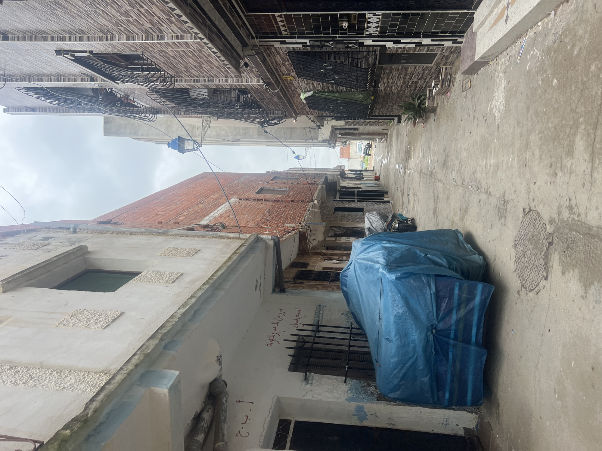 The image depicts a narrow alley in Martil, Morocco, as evidenced by the architecture and script. The alley is paved with concrete and appears damp, suggesting recent rain or washing. Buildings line both sides, exhibiting varied construction styles, including exposed brick, whitewashed facades, and decorative ironwork. A large object covered in a blue tarp is prominent on the right side of the alley. Electrical wires crisscross overhead, and a street lamp is visible in the distance. The sky is overcast, casting a muted light on the scene. Arabic script is visible on the wall of one building. In the distance, vehicles are parked.