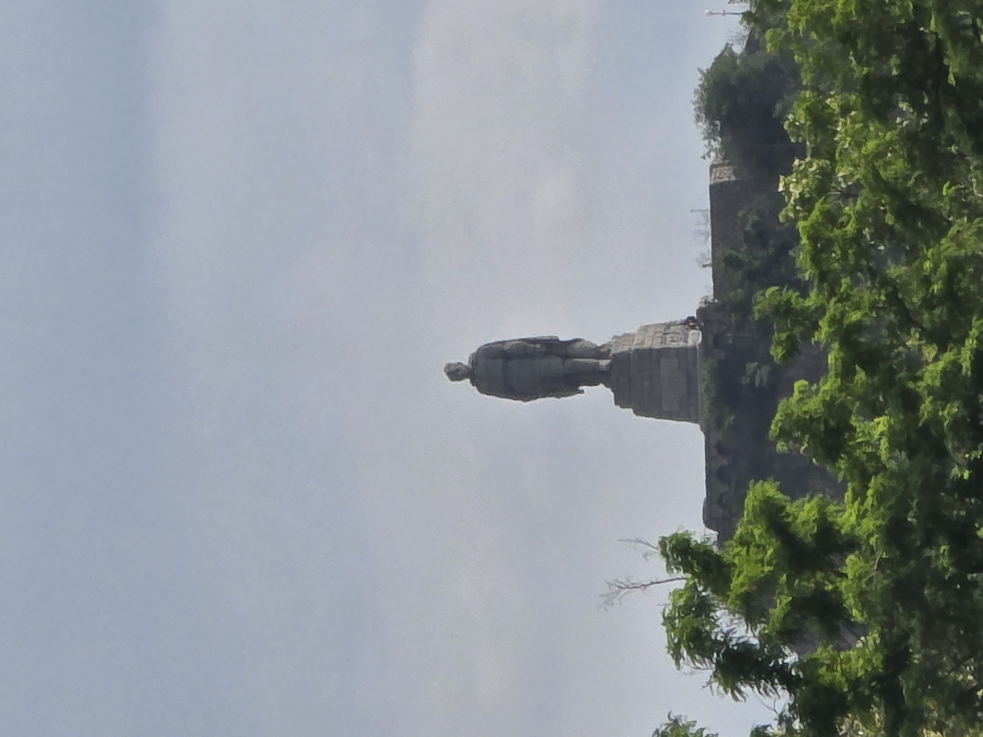 The image displays a prominent, towering statue of a standing male figure, set against a bright, hazy sky. The statue, crafted from a dark material resembling bronze or stone, depicts a man in traditional attire, holding what appears to be a staff or walking stick in his left hand. It rests on a substantial, multi-tiered stone base.
Adjacent to the statue's base, on the right side, there's a dark-colored structure, possibly part of the monument's platform or an associated building, showing some architectural details. Dense, vibrant green trees are visible in the foreground and to the right, partially obscuring the lower portions of the base and the background structure.
The scene is illuminated by daylight, with the sky appearing pale blue or light grey, indicating either an overcast condition or atmospheric haze. No specific activity is depicted, only the static presence of the monument. There is no visible text in the image. The overall impression is of a large, significant outdoor monument or landmark.
