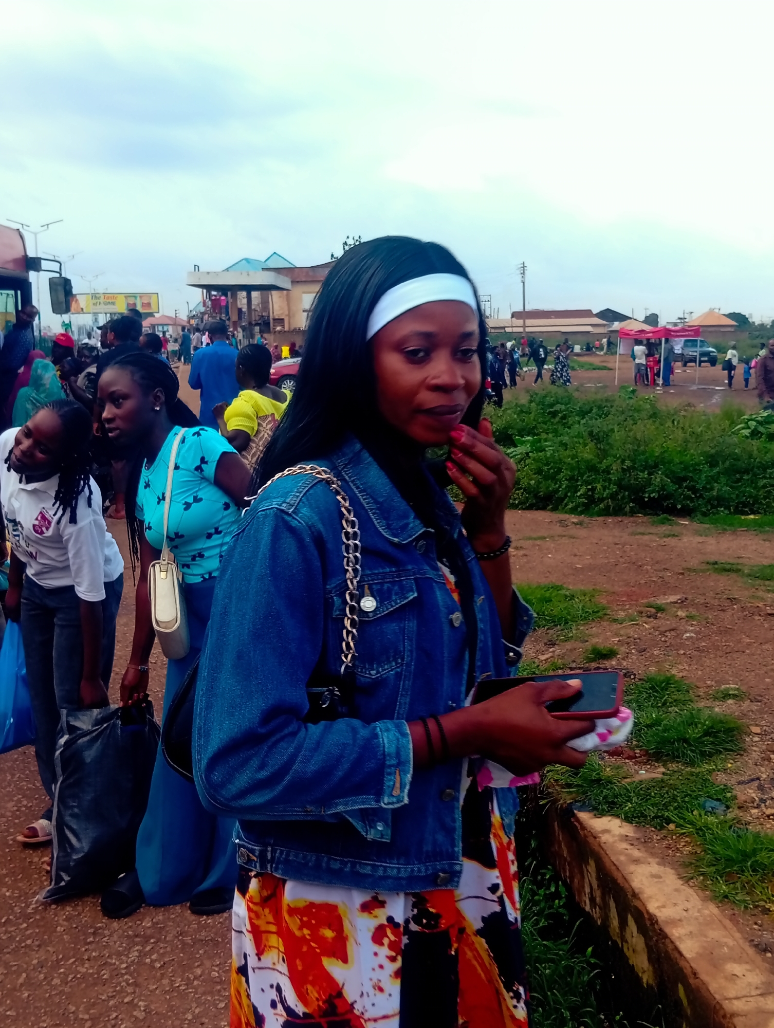 In Bukuru, Nigeria, a street scene unfolds under a cloudy sky. The foreground is dominated by a woman wearing a denim jacket and a vibrantly patterned skirt, holding a smartphone and a cloth. Behind her, other individuals are visible, including a woman in a turquoise shirt and a white handbag, and another in a white shirt. The background shows a bustling environment with a bus, market stalls, and a building with blue roofing, suggesting a commercial or transportation hub. The ground is a mix of dirt and gravel, with patches of greenery.