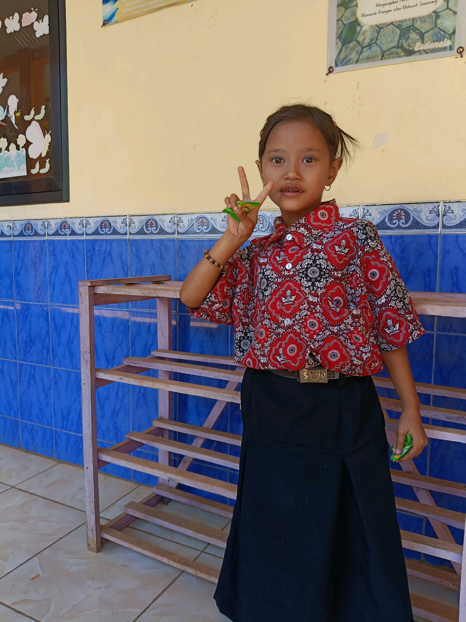 A young girl with dark hair pulled into a ponytail stands in front of a blue tiled wall and a wooden shelf. She is wearing a red and white patterned shirt and a black skirt. Her right hand is raised, and she is holding up two fingers in a peace sign. A small green object is visible in her left hand. The background shows a cream-colored wall with a poster at the top right and a framed picture with butterflies on the left. The floor is tiled in a beige and white pattern.
