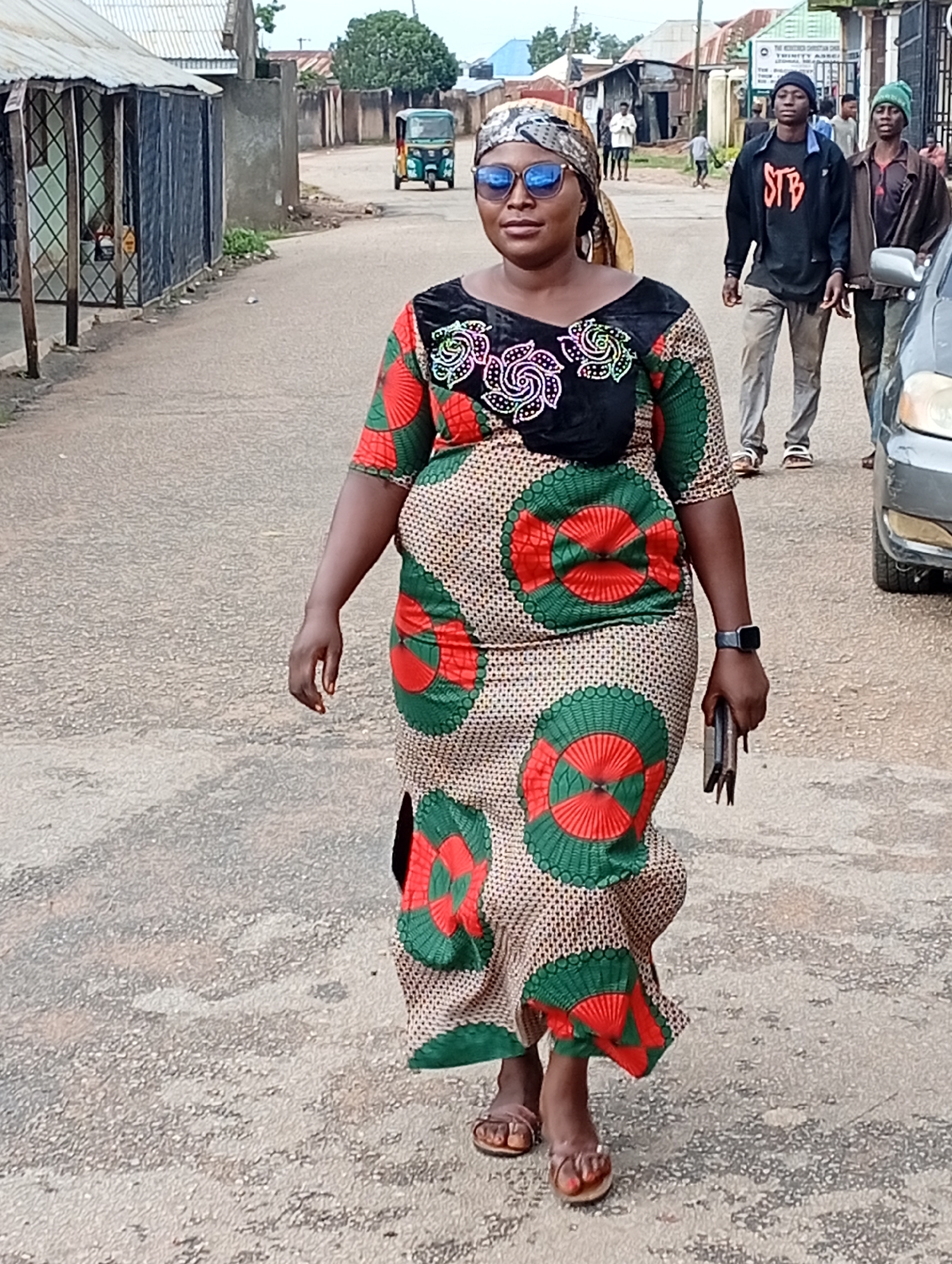 The image captures a street scene in Jos, Nigeria. In the foreground, a woman walks towards the camera, wearing a patterned dress and a headscarf. She has sunglasses on and is holding a phone in her right hand. Behind her, two young men walk away from the camera, and a three-wheeled vehicle is visible in the background. The street is paved and lined with buildings and shops. The overall atmosphere suggests a typical day in an urban environment.