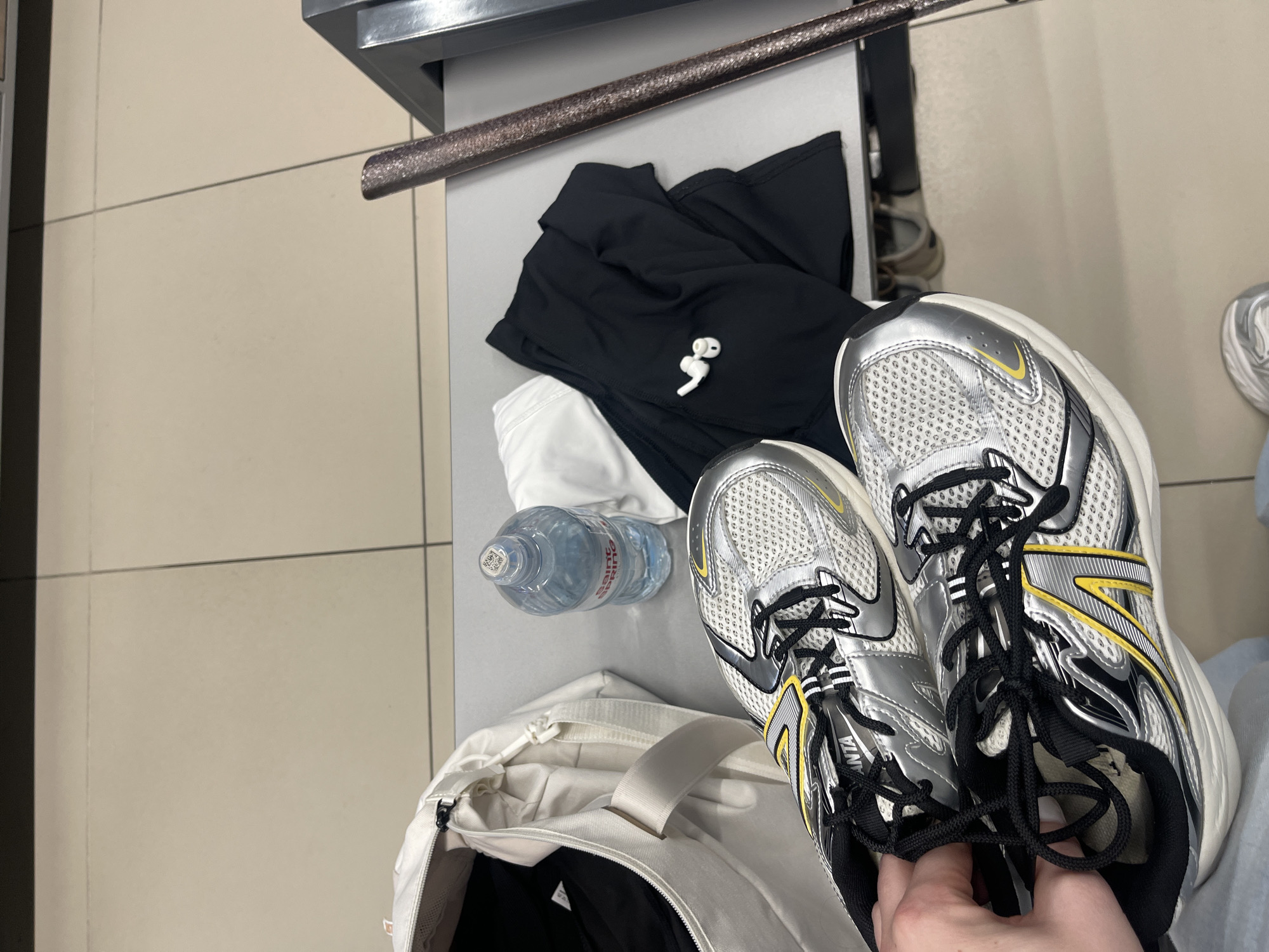 The image shows a pair of silver and yellow athletic shoes being held by a person's hand in the foreground. In the background, on a grey table, is a white duffel bag with black clothing inside, a bottle of water, and a pair of black athletic pants with white wireless earbuds placed on them. To the right of the table is a metallic object. The floor is tiled. The scene is likely in a public locker room or a similar facility, possibly at a sports venue. Without further context, the specific location within Minsk, Belarus cannot be determined.
