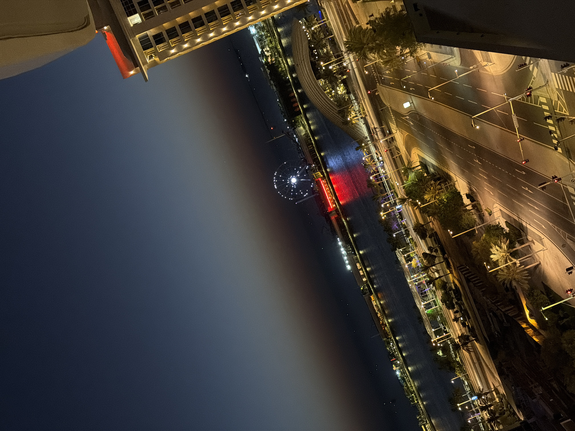 A high-angle, wide shot captures a nighttime scene of a city waterfront. The image is tilted, with a large building on the right side of the frame in the foreground. This building, illuminated by numerous lights, has a distinct red illuminated band near its top. Below the building and extending into the midground is a body of water reflecting the lights of the city. Across the water, a Ferris wheel with white lights is visible in the distance, along with other illuminated structures and a line of lights that appear to be a waterfront promenade or buildings. In the foreground, a road network with streetlights and traffic signals cuts through a landscape of palm trees and other greenery. The sky transitions from a deep blue at the top of the frame to a dark orange and then black towards the horizon, indicating either dusk or a clear night. No people are discernible in the image.