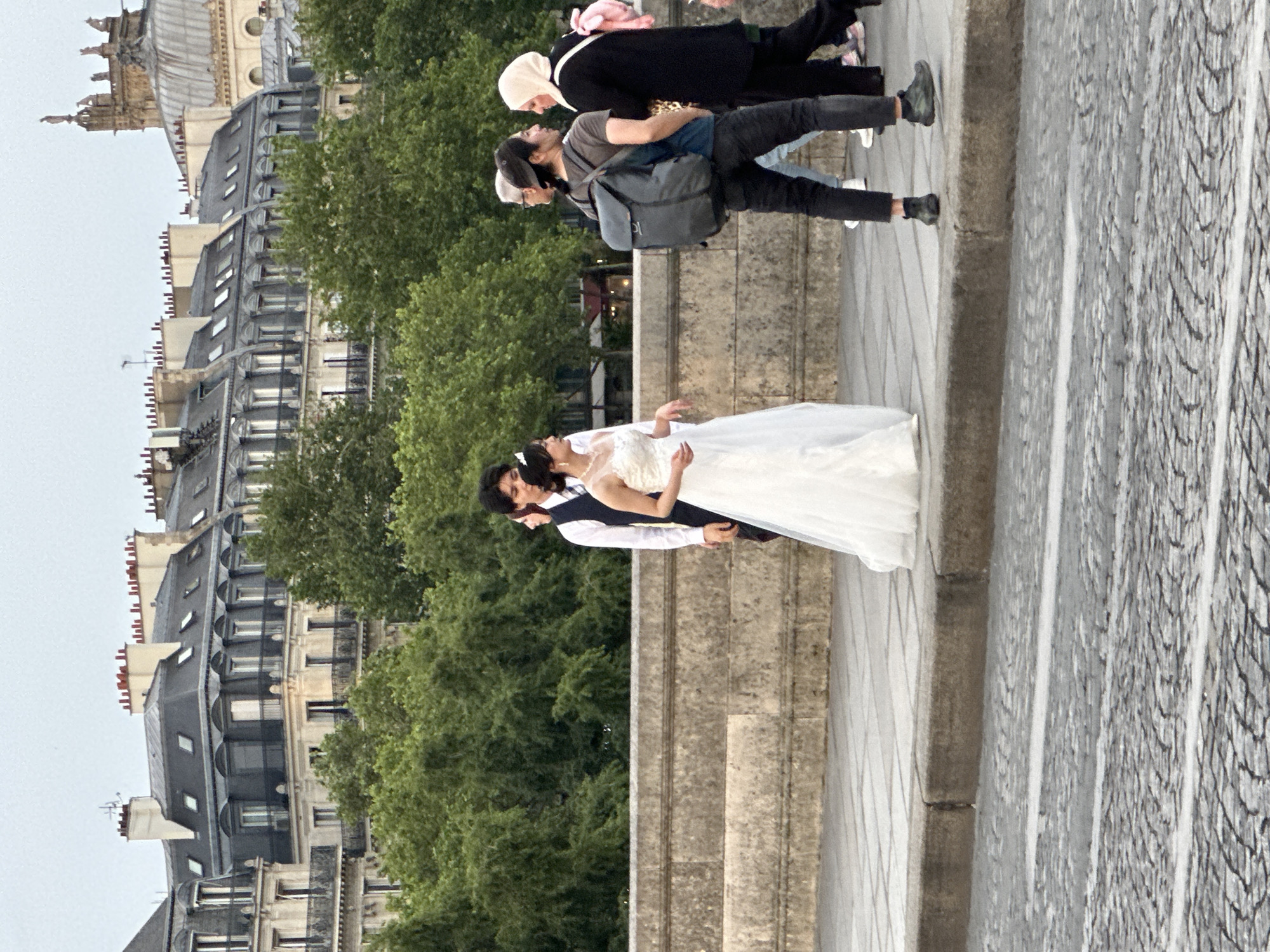 A wedding scene unfolds on a bridge in Paris, France. The bride and groom are embraced in the center. The bride wears a white gown, and the groom is in a dark suit. Pedestrians walk by behind them. Parisian architecture is in the background.