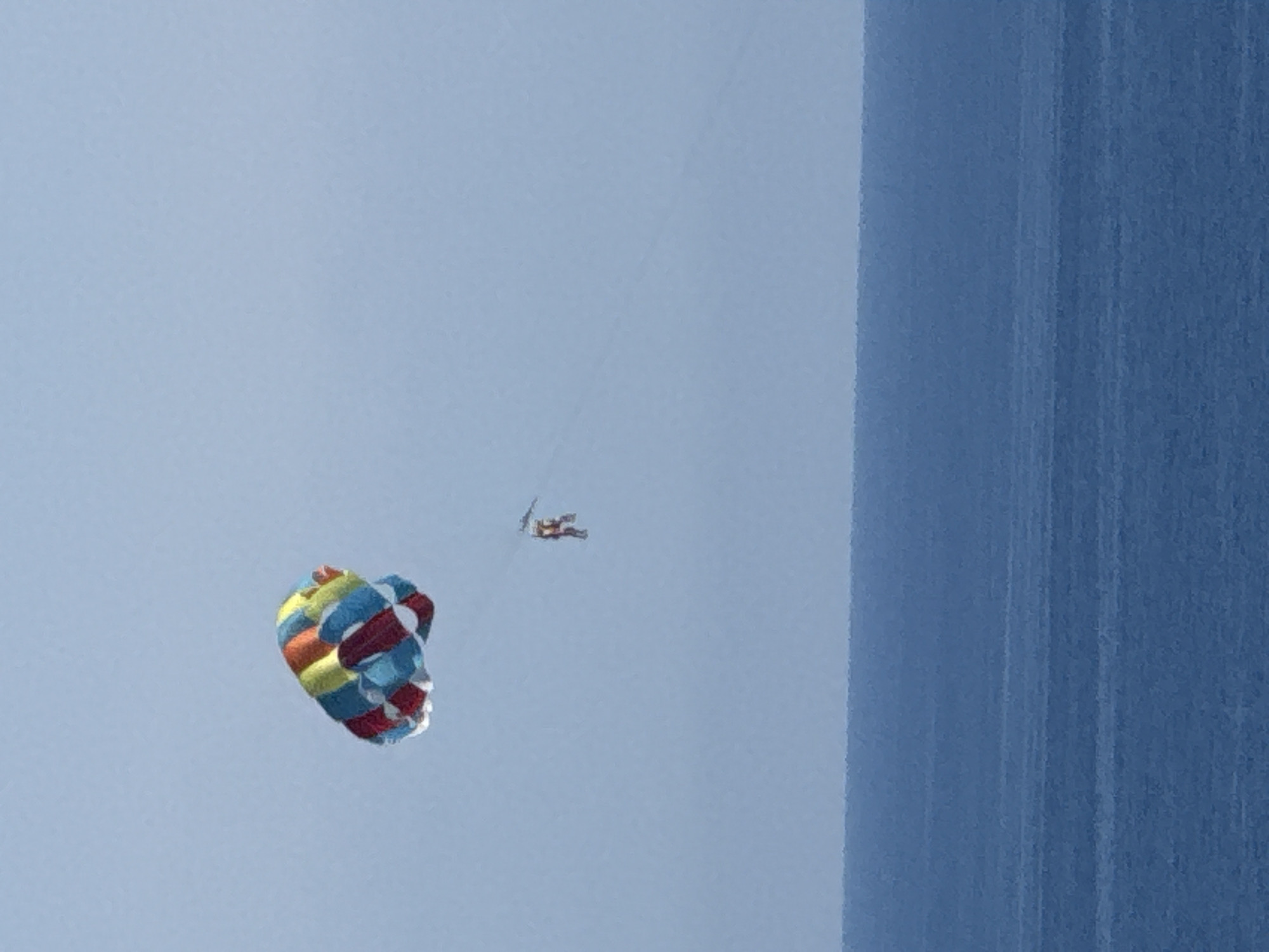 A paraglider is suspended in the air over the sea, towed by a boat, in the city of Boreti, Montenegro. The paraglider is colorful, with sections of red, yellow, blue, and white. The person is seated in a harness attached to the paraglider. The sea is a deep blue, with gentle ripples on the surface. The sky is a pale blue. The image is taken from a distance, capturing the vastness of the sky and sea.