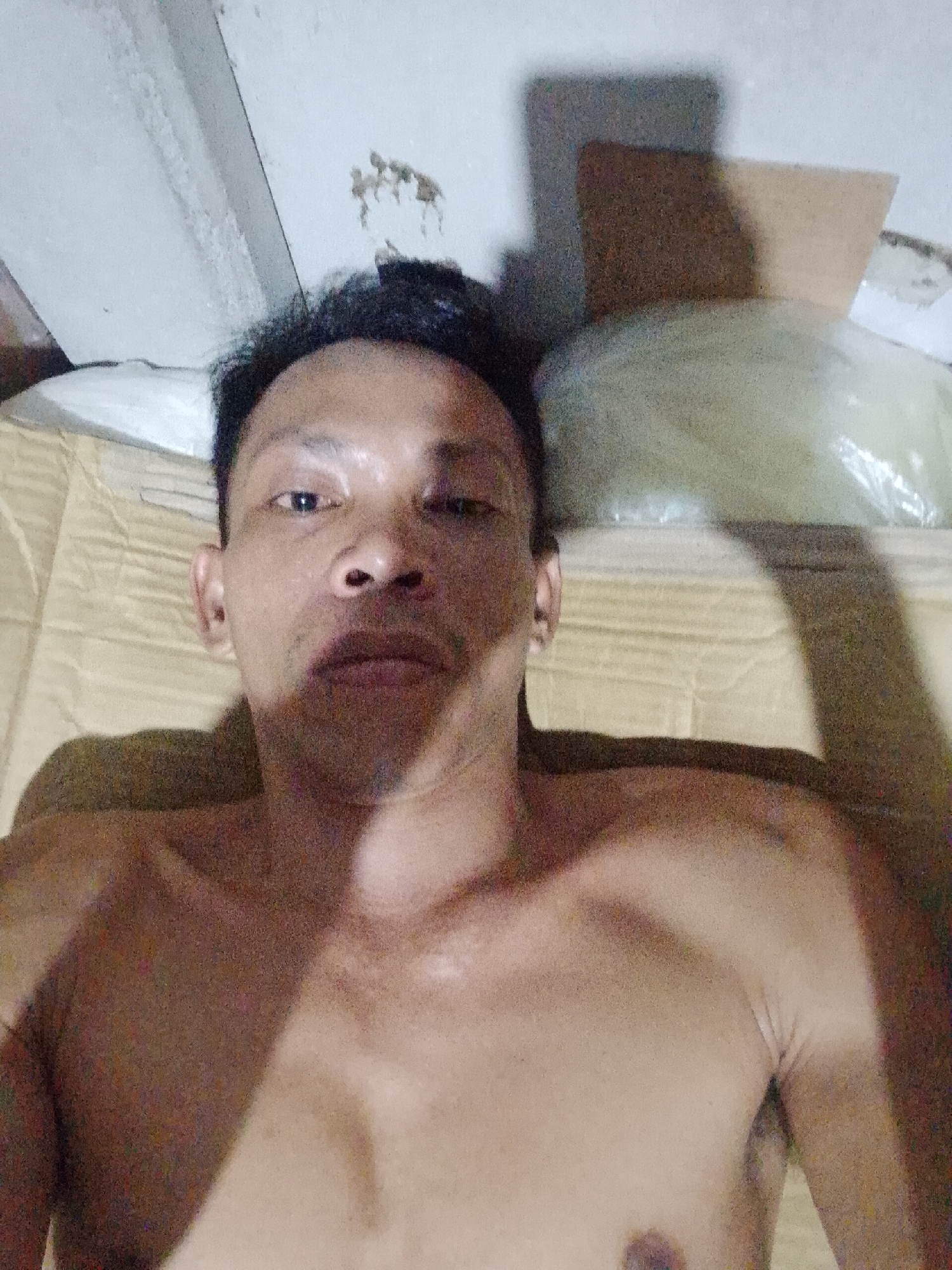 The image depicts a shirtless man lying on a tan, patterned surface, possibly a mattress or futon. He has short, dark hair and appears to be middle-aged. Above him, a white ceiling with stains and exposed concrete is visible. In the upper right corner, there is a plastic-covered object, along with a piece of cardboard casting a shadow. The photograph appears to have been taken indoors in Bukit Ketok, Indonesia.