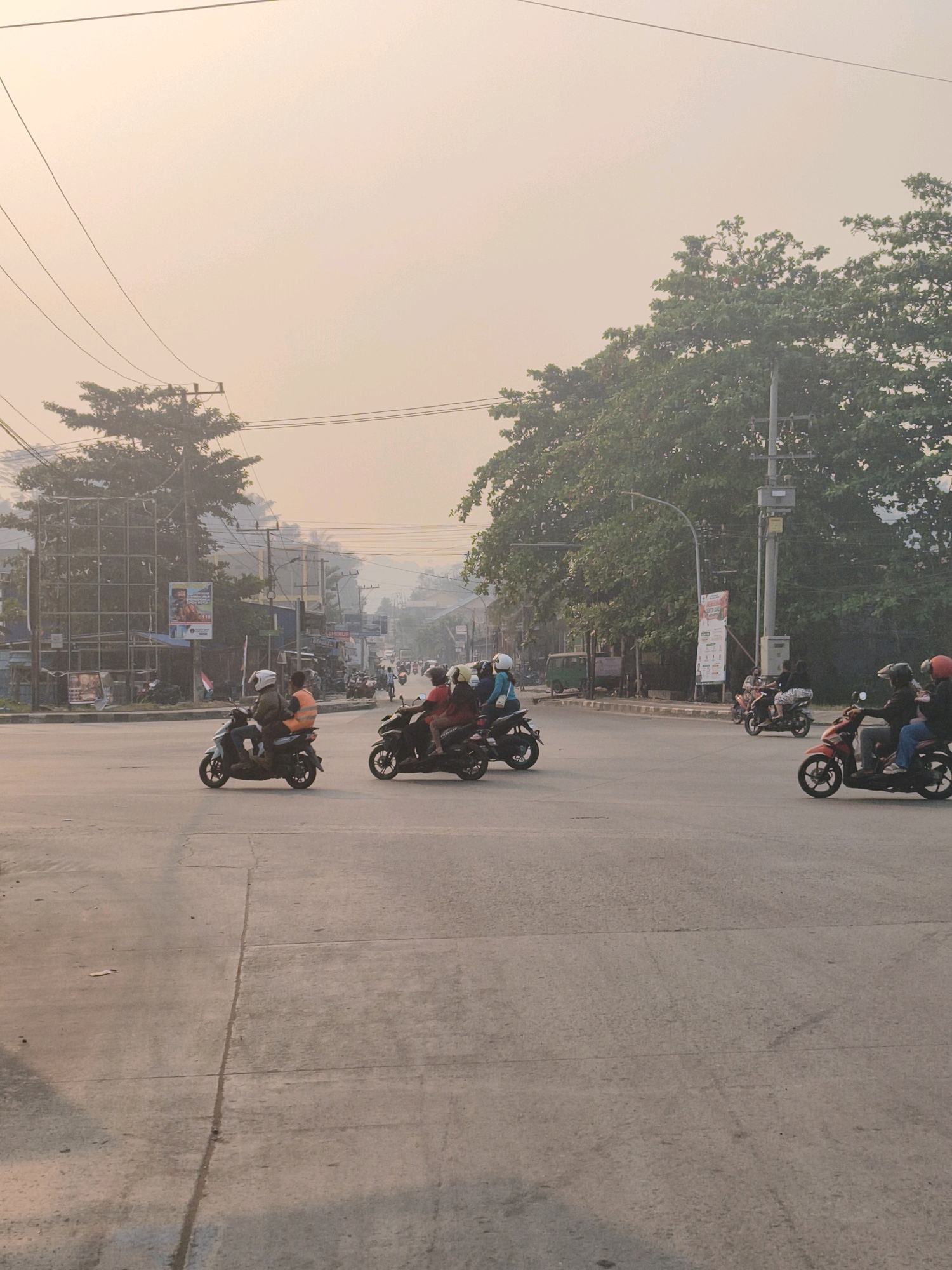 This image captures a street intersection in Samarinda, Indonesia, likely during the morning or late afternoon given the soft, hazy light. The scene is dominated by a wide, paved road with visible expansion joints, indicating a potentially busy urban thoroughfare. A number of people are traveling on motorcycles, which are the primary mode of transportation depicted.
In the foreground, to the left, a white scooter carries two passengers; the rider wears a helmet and a light-colored jacket, while the passenger behind him sports a bright orange vest and a helmet. To the right of this scooter, a black motorcycle is also carrying two individuals, both wearing helmets, with the rear passenger in a red top. Further into the intersection, a red scooter with two helmeted riders is moving to the right. In the middle ground, several other motorcycles with riders are visible, suggesting moderate traffic flow.
The environment includes significant greenery, with large, leafy trees lining the right side of the road, their branches extending over the street. On the left side, a billboard structure stands, and buildings and various signs are visible in the background, interspersed with utility poles and overhead wires, characteristic of an urban setting in Indonesia. The sky is overcast with a light, muted tone, contributing to the diffused lighting. The overall impression is of a typical street scene in Samarinda, with everyday activities of commuting on motorcycles set against a backdrop of urban infrastructure and abundant vegetation.