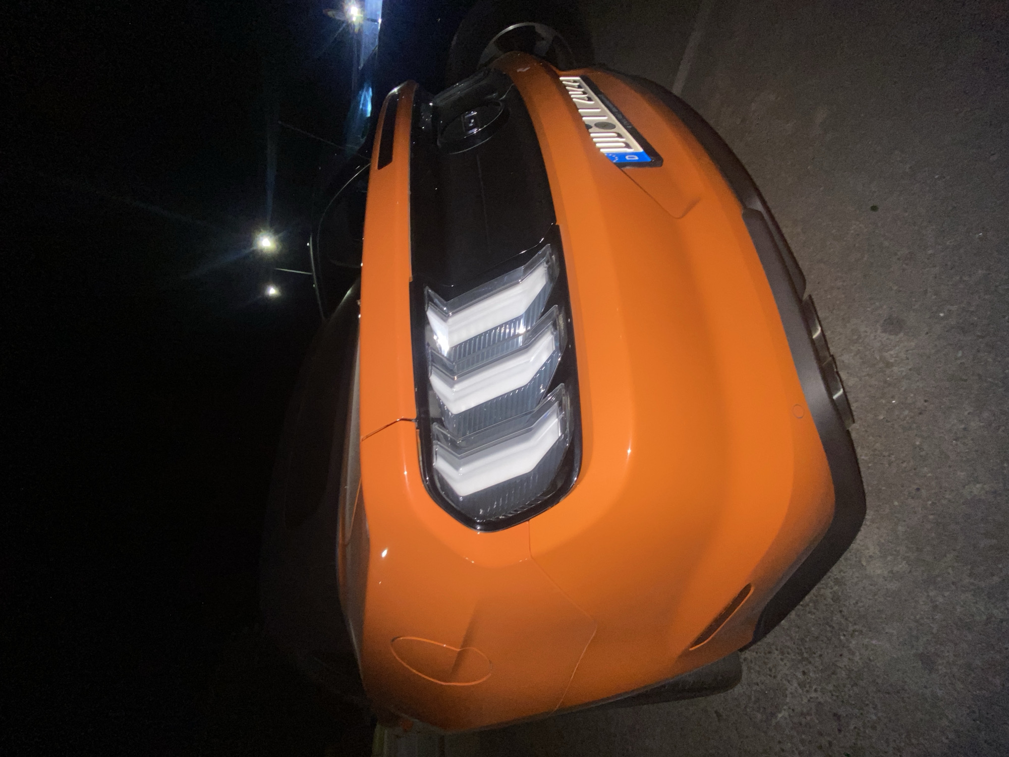 The image shows the front of an orange Corvette C8. The car is parked at night, likely in an urban environment, and possibly the city of Bar, Montenegro, given the prompt. The headlights are on. The license plate is partially visible, though unreadable. The car is parked on asphalt.