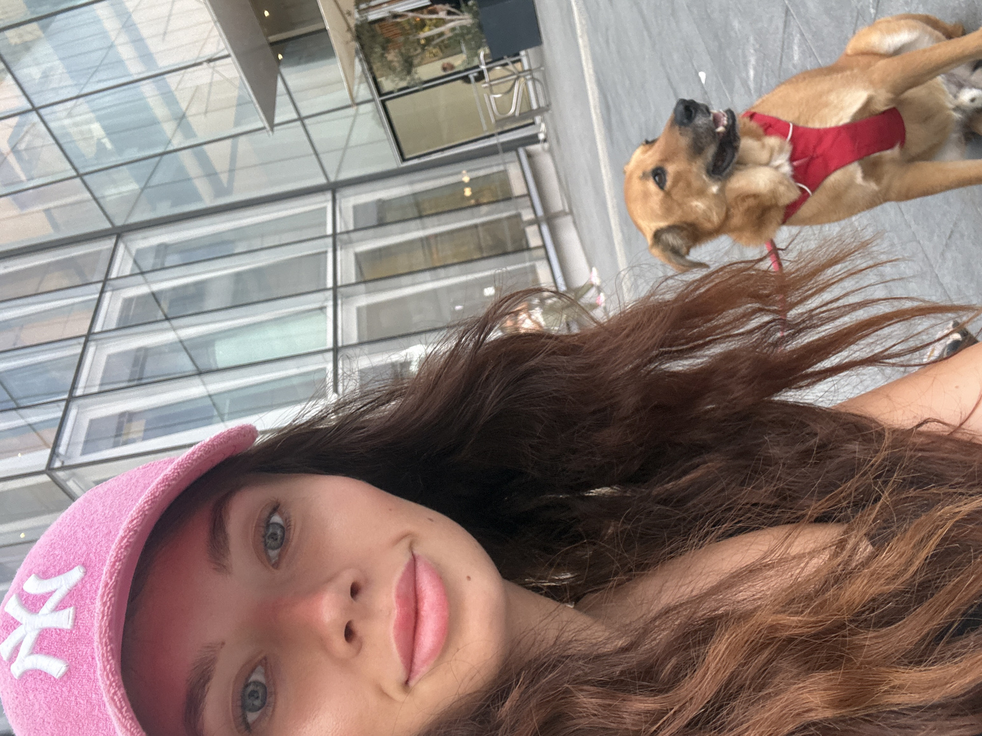The image is a selfie taken by a young woman wearing a pink baseball cap with a white "NY" logo. She has fair skin, brown hair, and blue eyes. Her lips are slightly parted in a smile. Behind her is a tan-colored dog wearing a red harness. The dog is looking up and to the right, seemingly at something or someone beyond the frame. The background reveals a modern building with large glass windows. Based on the architectural style and the visible signage (not clear enough to decipher), it is possible this image was taken in an urban setting resembling the city of Tel Aviv, Israel. The overall composition suggests a casual, outdoor moment captured during a walk or outing in the city.