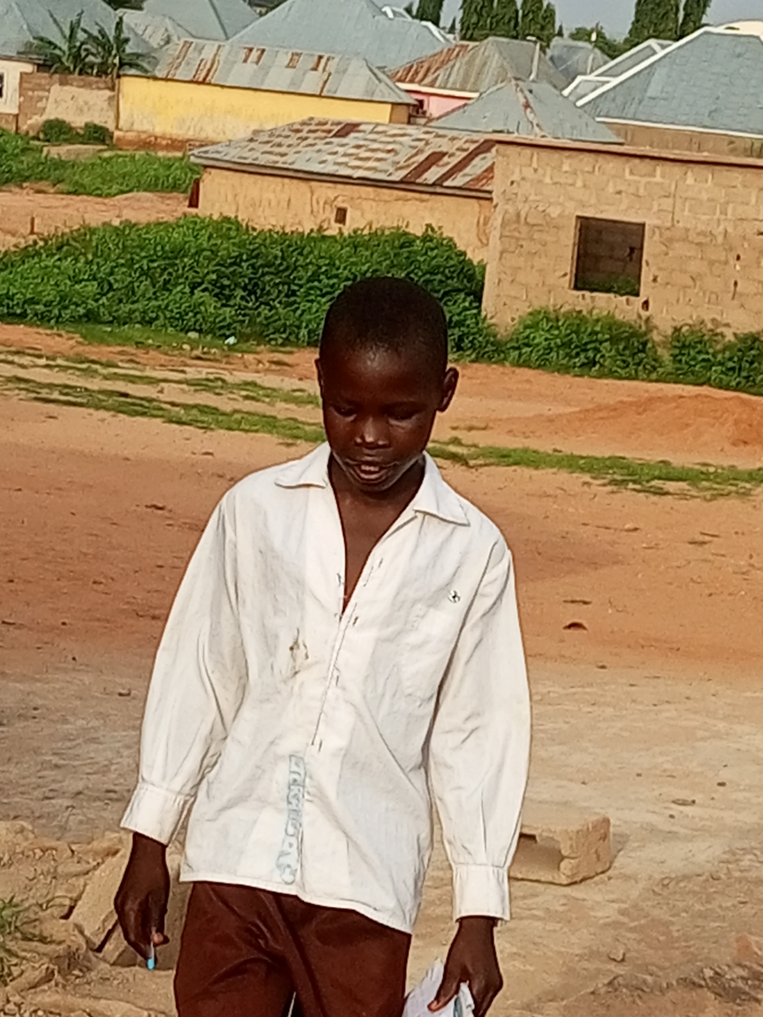 In Tumfure, Nigeria, a young boy stands outside a building, wearing a white collared shirt and brown trousers. He is holding a small blue object in his right hand, possibly a pill or a small toy. The background features several buildings with corrugated metal roofs, typical of the local architecture. The ground is sandy and uneven, with some patches of green vegetation. The overall scene depicts a rural or semi-urban environment.