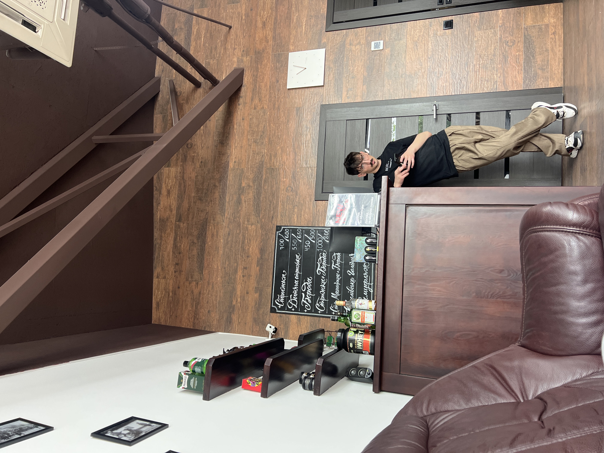A slightly rotated interior scene features a person lying on a brown wooden cabinet with a black chalkboard menu attached, near a dark brown leather armchair. The setting includes a stairwell in the background, and the flooring and wall have a matching brown wood pattern. A white clock is mounted on the wall.
The person is wearing a black t-shirt and beige pants, with white and black sneakers. They are positioned horizontally on top of the cabinet, with their head near the edge and feet toward the back.
The chalkboard menu lists various services in Cyrillic, including haircuts and beard treatments, along with prices in what appears to be Ukrainian currency. Below the menu, there are shelves holding bottles and products.
In the foreground, there are black-framed photographs on a white surface. The stairwell is to the left, ascending into the upper part of the scene.