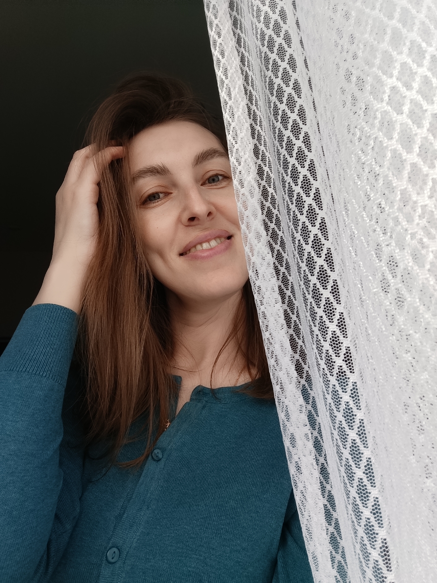 The image portrays a woman with long, reddish-brown hair, smiling warmly at the camera. She appears to be in her 20s or 30s and has blue eyes. Her right hand is raised, gently touching her hair on the side of her head. She is wearing a teal or dark green long-sleeved cardigan with buttons down the front.
The setting appears to be indoors, possibly near a window, with a dark, indistinct background. A white, sheer curtain or netting with a honeycomb-like pattern occupies a significant portion of the right side of the frame, partially obscuring the woman's face and body. Natural light seems to be softly illuminating her from the side where the curtain is.
The woman's expression is joyful and relaxed, suggesting a pleasant mood. She seems to be playfully peeking from behind the curtain, posing for the photograph. There is no visible text in the image. This scene is set in Chaikovsky, Russia.