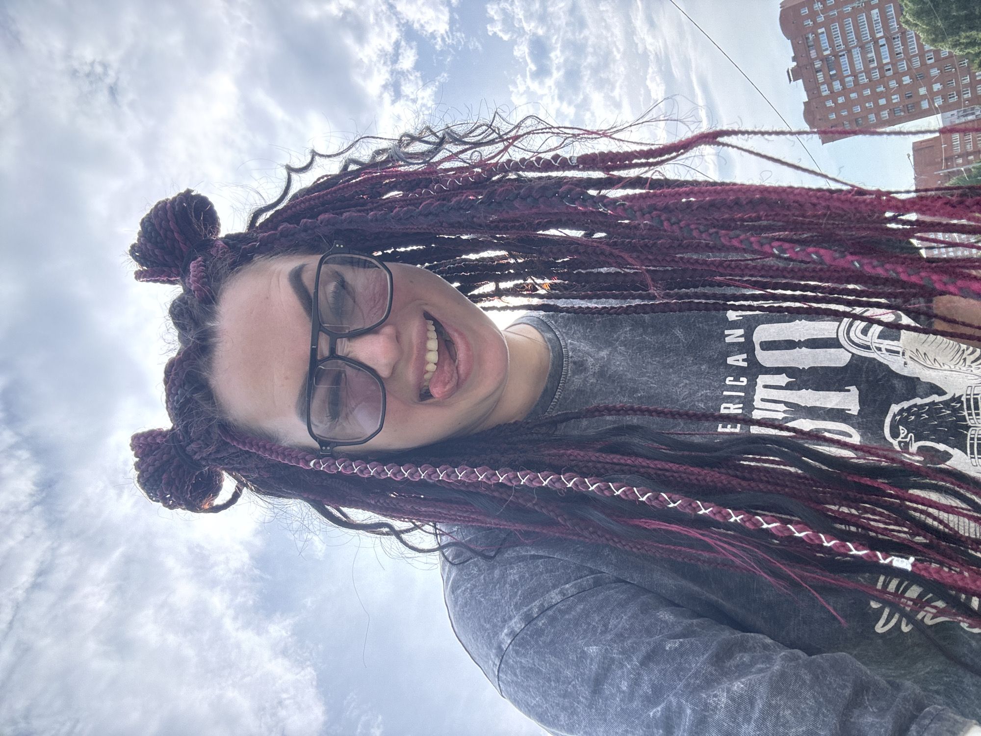 A young woman with dark hair styled in burgundy braids that are tied into two buns on top of her head, is sticking her tongue out at the camera. She is wearing black-rimmed glasses and a gray t-shirt with a graphic design featuring an eagle and text that reads "AMERICAN THUNDER". The photo is taken from a low angle against a cloudy sky. In the background, there is a brick apartment building and some trees, suggesting an urban setting, possibly in Tolyatti, Russia.