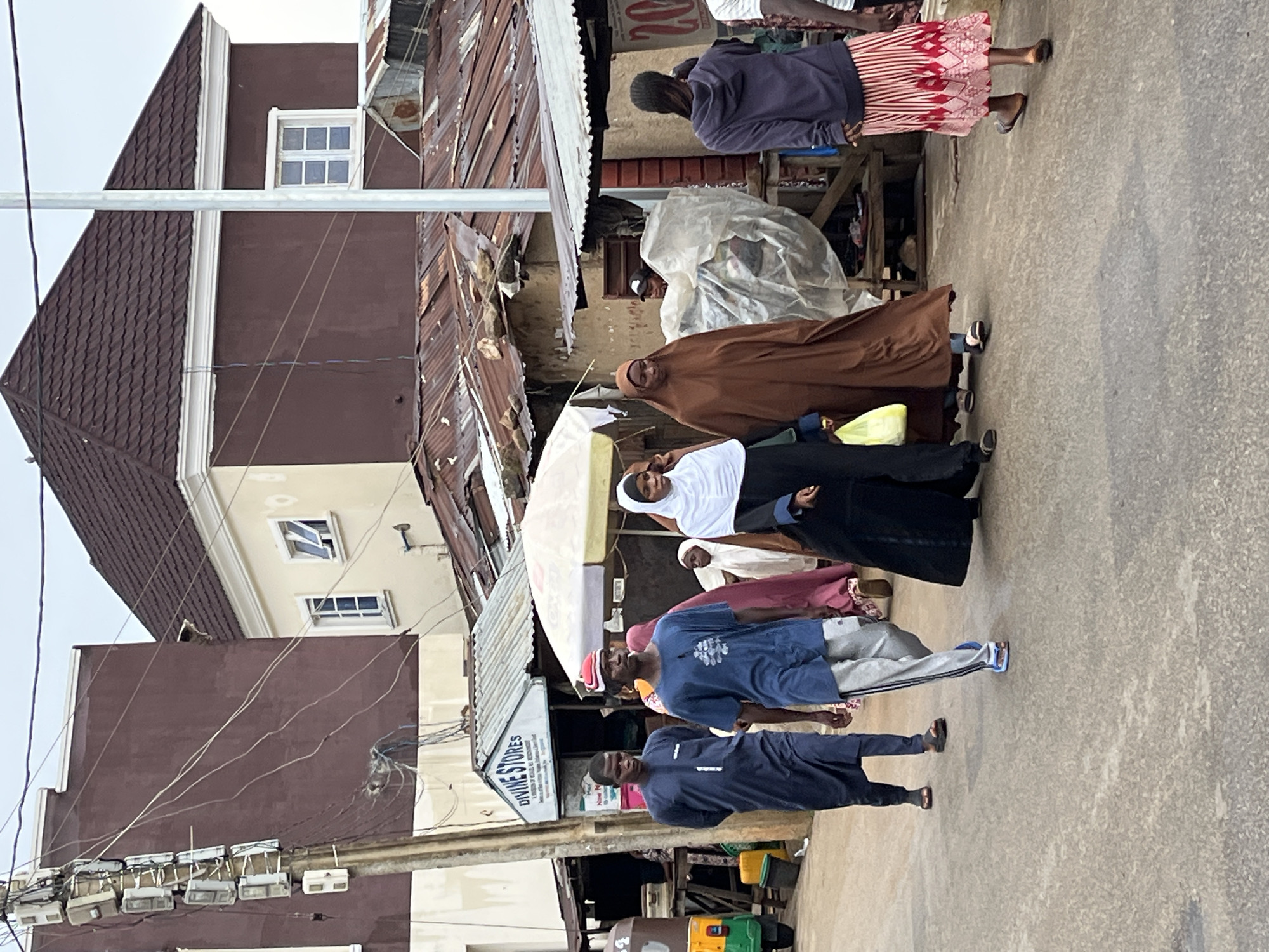 The image shows a street scene in Jos, Nigeria. A group of people are walking along a street with buildings on either side. The buildings vary in construction style, with some appearing older and more makeshift, while others look more modern. Utility poles and wires are visible overhead. The people are wearing a mix of traditional and casual clothing. Some women are wearing hijabs or full-body coverings, while others are dressed in more modern attire. A few men are wearing traditional robes. The street appears to be paved and is somewhat crowded with pedestrians. There are shops and stalls along the sides of the street, with signs visible above them. A tricycle taxi (Keke Napep) is also present. The scene captures the everyday life and diverse character of a street in Jos.