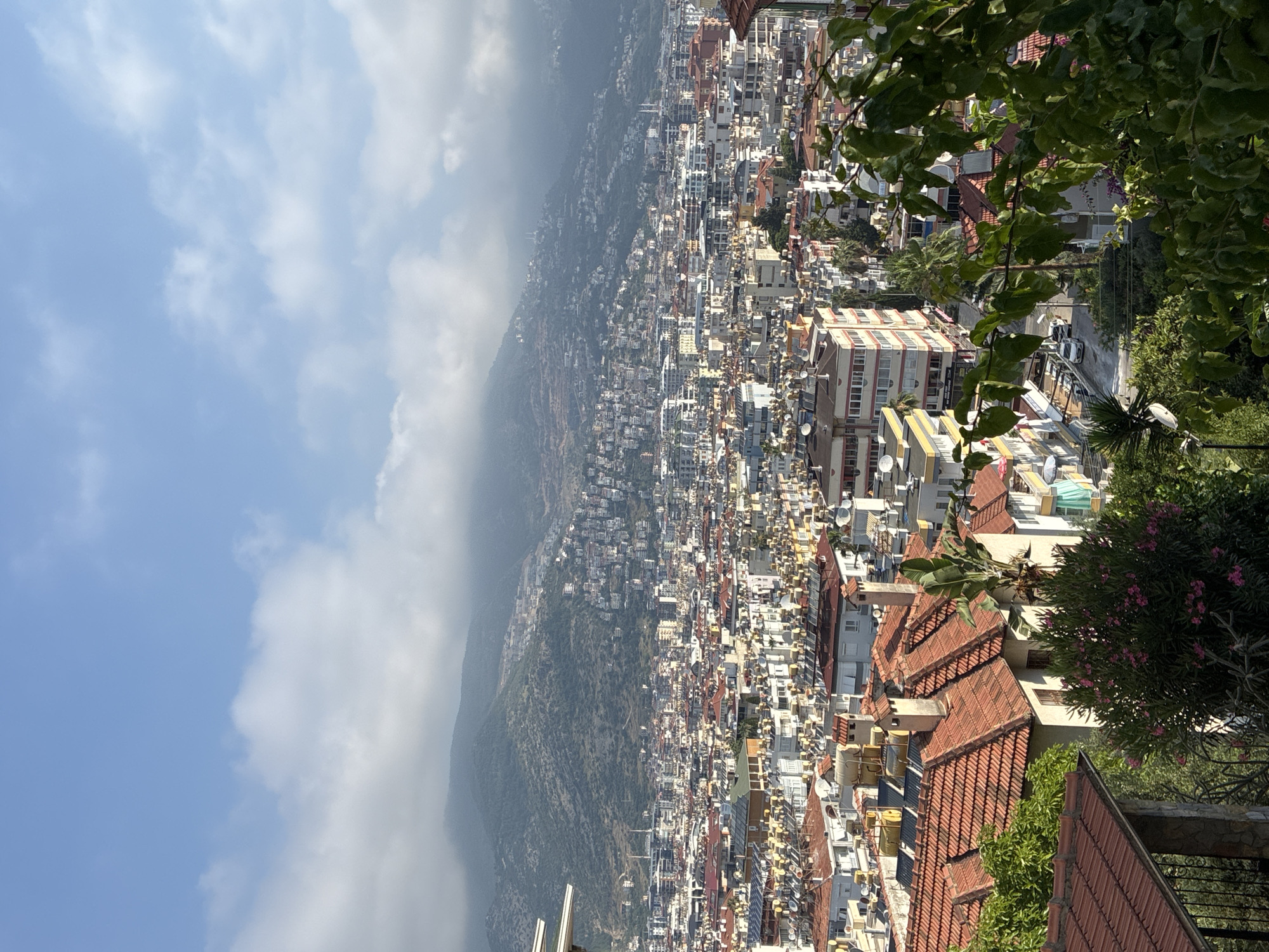 This is an elevated, wide shot of a dense urban landscape in Alanya, Turkey, nestled against a backdrop of green, mountainous terrain partially obscured by clouds. The foreground features a residential building with a red-tiled roof, partially covered by lush green foliage and flowering bushes with pink blossoms. This building is situated on a hillside, with a stone and metal fence visible to its left.
Beyond this foreground, the city unfolds. Numerous residential buildings, primarily with red-tiled roofs and varying architectural styles, dominate the mid-ground. Many of these buildings are equipped with solar water heaters, evidenced by the numerous cylindrical tanks on their rooftops. The streets between the buildings appear narrow, with some parked cars visible.
In the background, the city extends up the slopes of the mountains. The mountains themselves are covered in a mix of vegetation and scattered buildings, with more distant peaks fading into a hazy, cloudy atmosphere. The sky above is a clear blue, with scattered white clouds adding depth and texture to the scene. There are no people clearly visible in the frame. The overall impression is one of a sprawling Mediterranean coastal city built on hilly terrain.