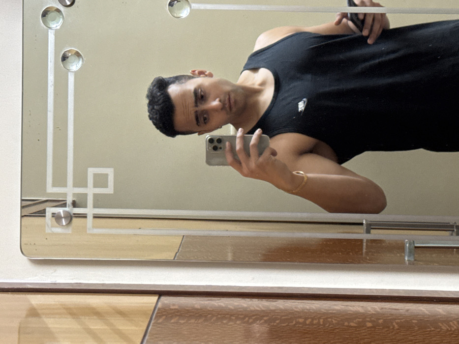 A man in a black tank top takes a selfie in a mirror. He holds a smartphone, likely capturing his reflection. The mirror features a geometric design at its corners. The background includes a wall and a section of a wooden surface, possibly a table or dresser. The composition suggests a casual, self-portrait style.