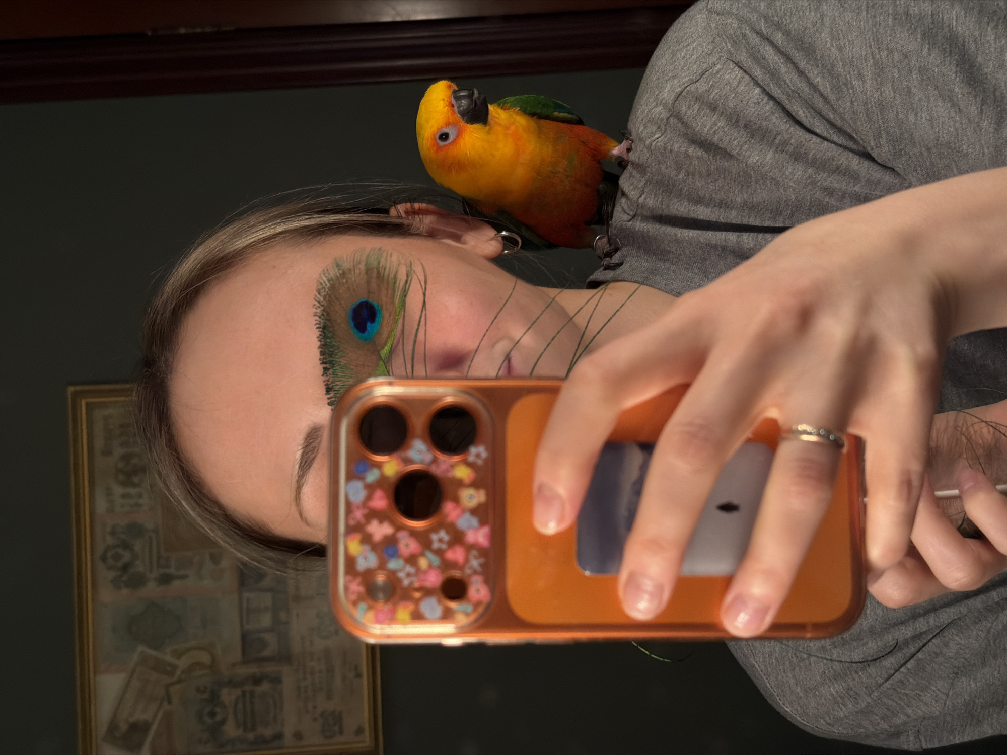A woman is taking a selfie with a cell phone in an orange case. She is wearing a grey shirt, and a parrot is perched on her shoulder. A peacock feather is attached to her forehead. The reflection in the phone case shows a patterned picture frame on the wall behind her. The image does not contain information about Khimki, Russia.
