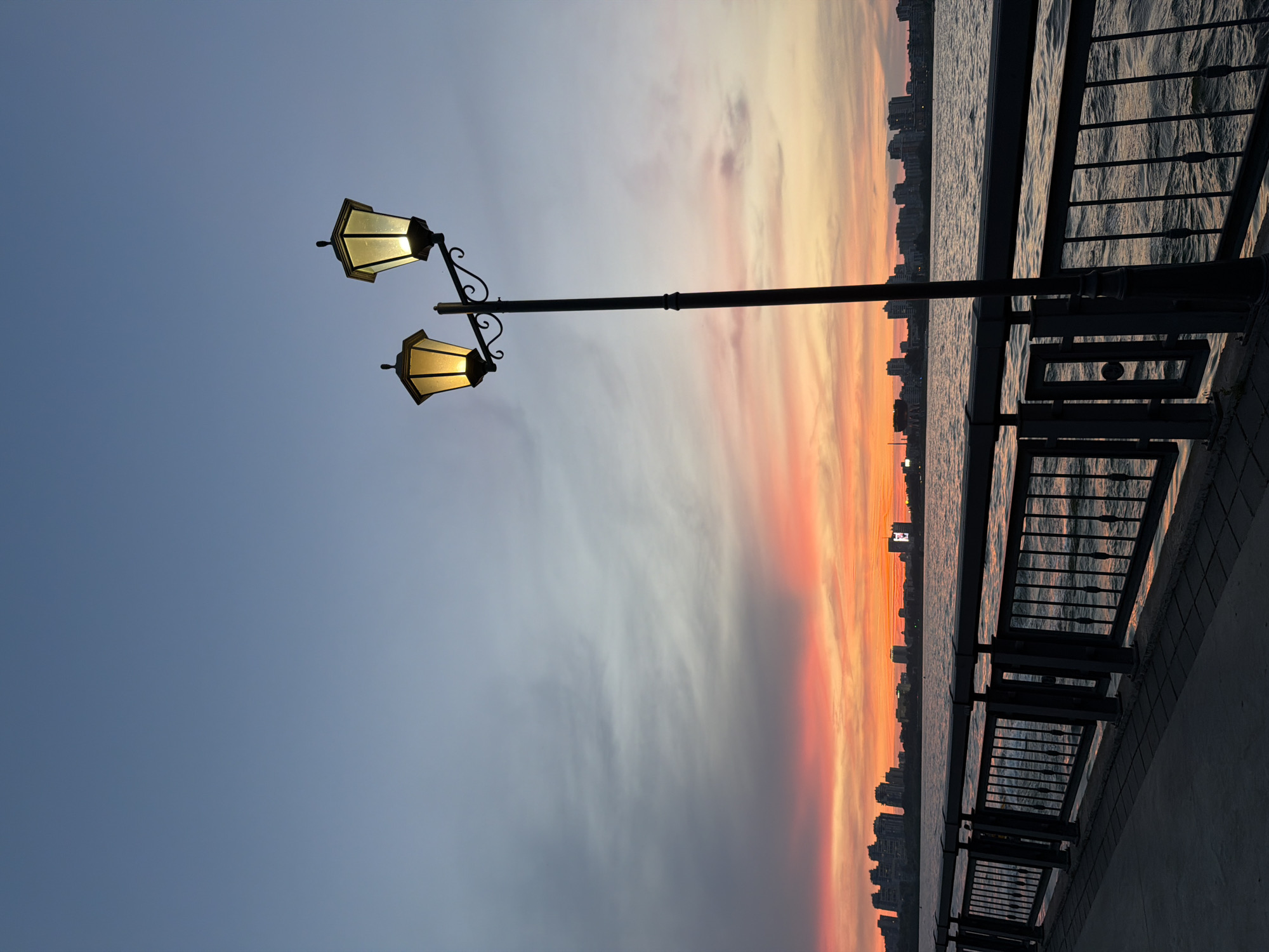 An outdoor vertical shot captures a waterfront promenade at dusk. A lamppost with two glowing lanterns stands in the foreground, casting a warm light against the twilight sky. The sky transitions from a clear blue at the top to shades of pink and orange near the horizon, indicating sunset. Wispy clouds are scattered across the sky. A dark metal railing separates the promenade from a body of water, likely the Volga River, which reflects the colors of the sky. In the background, a silhouetted city skyline with various buildings is visible. The promenade itself is paved with concrete tiles. No people are present in the image. The scene is tranquil, depicting a serene evening in Kazan, Russia.