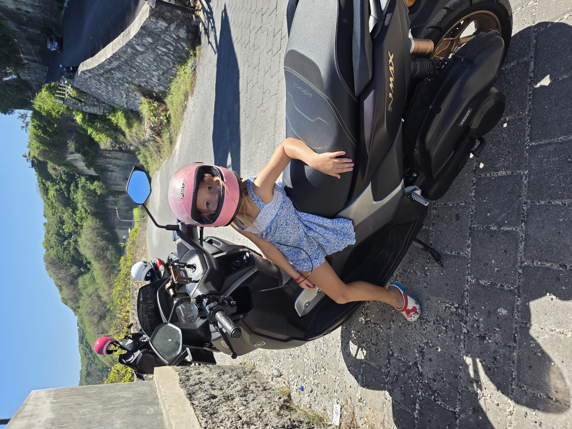 A young girl with fair skin and blonde hair, wearing a pink helmet and a blue floral dress, is seated on a black Yamaha NMAX scooter. Her right hand rests on the seat of the scooter, and her left arm extends forward towards the handlebars. She is wearing red and white clogs on her feet.
The scooter is parked on a paved surface, which appears to be part of a road or parking area. There is another scooter parked behind the one the girl is seated on, also with a pink helmet. A white helmet is also visible.
In the background, there is a green hillside and a clear blue sky. A grey stone wall runs along the edge of the road.
The location is Kuta Selatan, Indonesia.