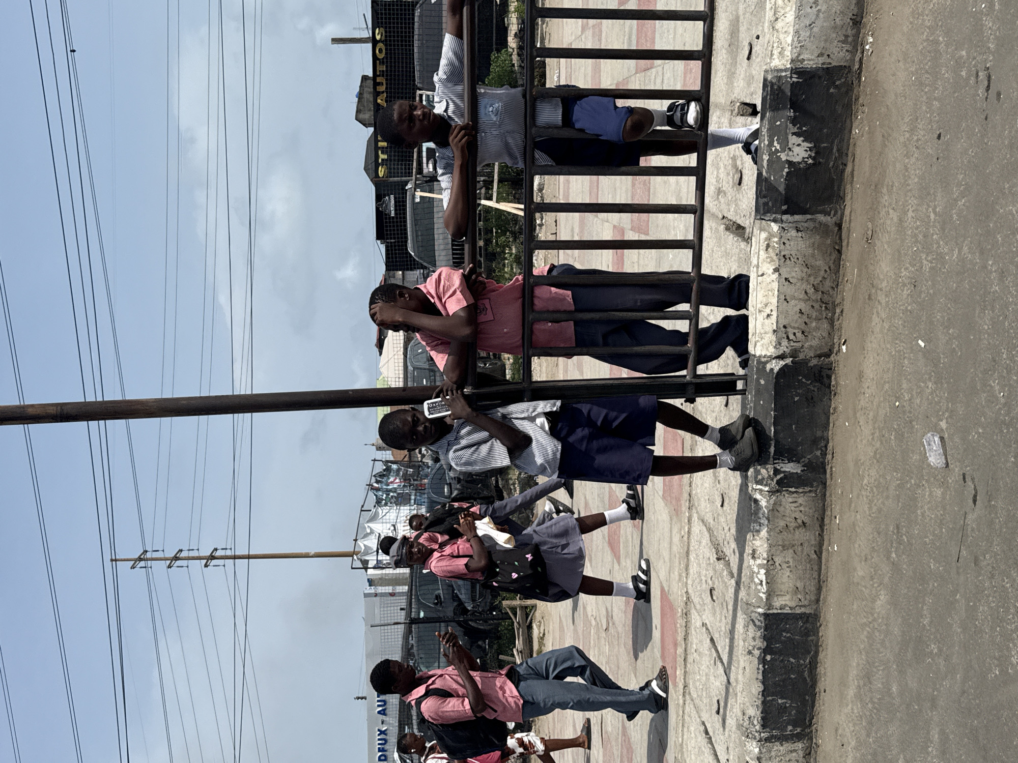 The image shows a group of schoolchildren in Igboefon, Nigeria, dressed in school uniforms and standing along a sidewalk next to a black metal fence. Some of the children are holding or using cell phones. A few of the kids are touching the fence while they are standing beside it. The background includes utility poles with wires, buildings, and signage, including a sign that includes the word "AUTOS". The weather appears to be sunny. The sidewalk has a black-and-white curb.
