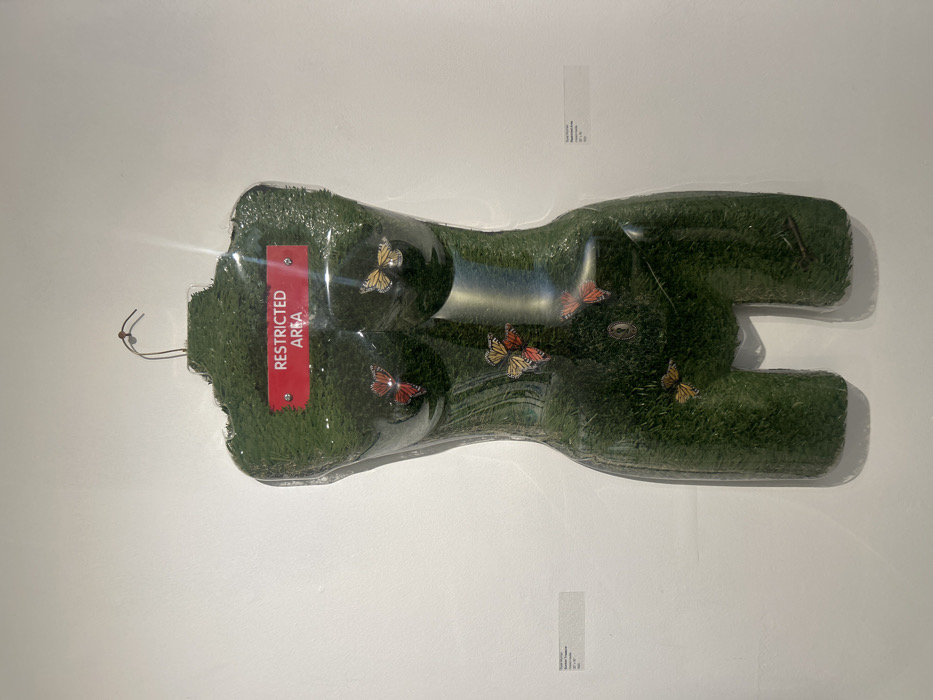 An artificial green lawn shaped like a woman's torso and legs is encased in clear plastic. A red rectangular sign reading "RESTRICTED AREA" is affixed near the torso's top. Decorative butterflies in orange, yellow, and red are scattered on the artificial grass. The artwork is mounted on a white wall, with an additional white label visible beneath the object.