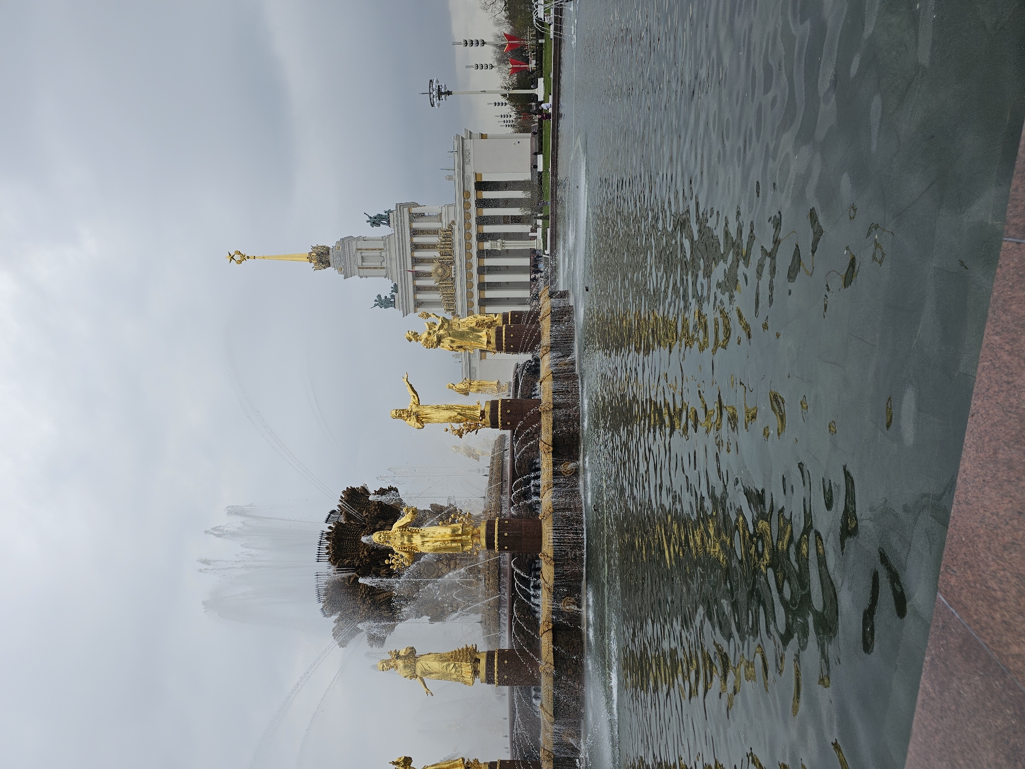 This media file captures a wide view of the iconic Friendship of Nations Fountain and the Central Pavilion at VDNKh (Exhibition of Achievements of National Economy) in Moscow, Russia. The image is oriented sideways, but depicts a grandeur outdoor scene on an overcast day.
In the foreground and midground, the magnificent Friendship of Nations Fountain dominates, featuring numerous golden statues standing around its multi-tiered structure. These allegorical figures, perhaps representing the former Soviet republics, are bathed in a soft golden hue. Water vigorously spouts from various points of the fountain, creating white splashes and streams that fall into the basin. To the right, a large, calm body of water (likely part of the fountain complex or a canal) reflects the muted sky and hints of the golden statues.
In the background, the grand Central Pavilion stands prominently. It is a classical-style building with a white facade, adorned with tall columns and intricate golden detailing. A towering golden spire, topped with a distinctive star, rises majestically from its center. Smaller golden statues are also visible along the pavilion's roofline. People can be seen as small figures near the building and along the fountain, suggesting visitors exploring the area. Further in the background, some trees and tall decorative light fixtures or poles with red banners are visible.
The overall atmosphere suggests a daytime visit, likely in spring or autumn given the muted light and lack of vibrant green foliage. The sky is largely overcast, casting a soft, diffuse light over the scene. There is no visible text in the image.