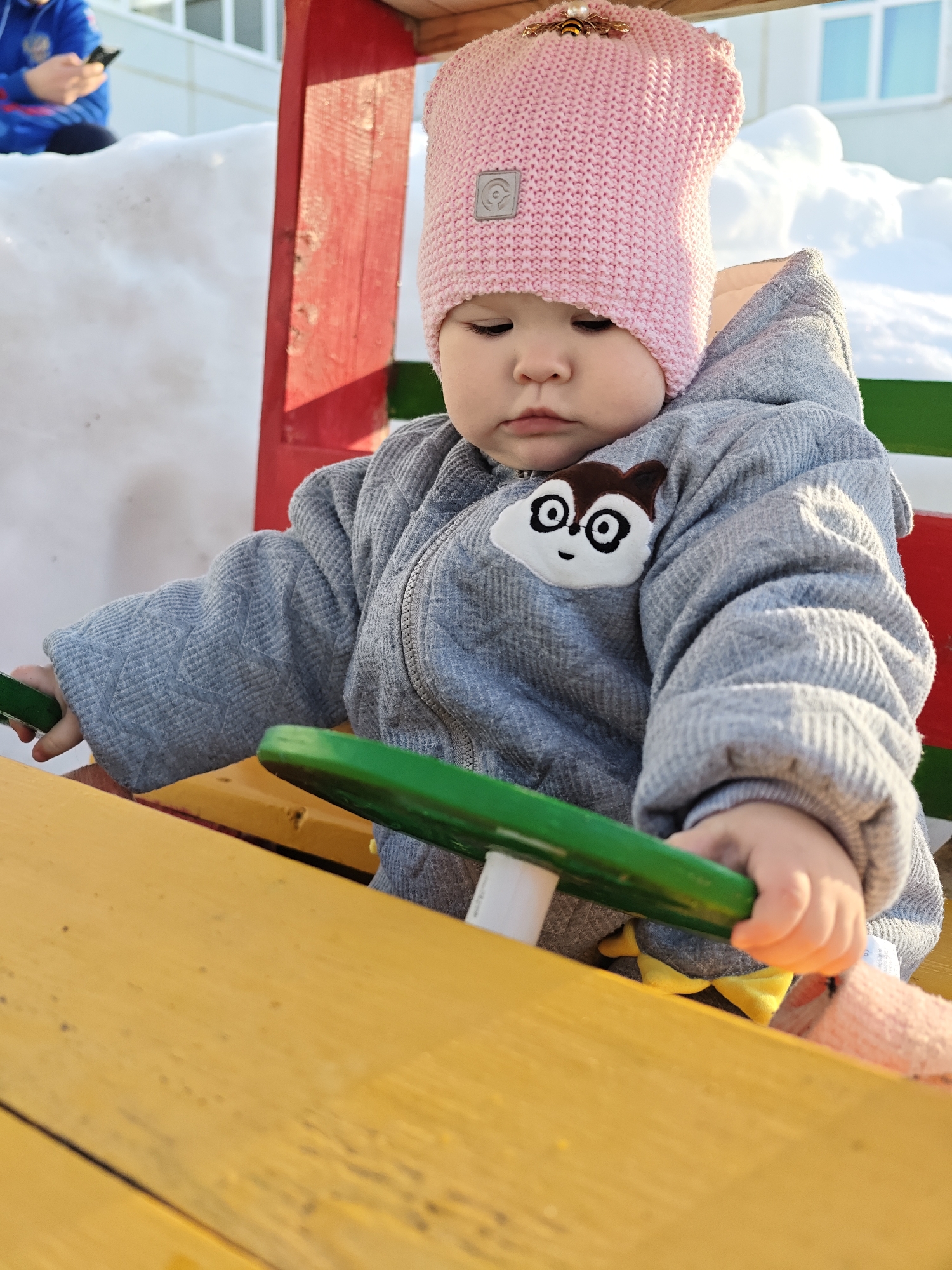 An outdoor scene in Chaikovsky, Russia, depicts a young child seated within a yellow wooden play structure, holding a green plastic steering wheel. The child wears a light pink knitted hat with a gold-colored bee ornament featuring a pearl-like body, and a grey quilted jacket with a white and brown cartoon raccoon appliqué on the left chest. The child's hands grip the steering wheel, and the gaze is directed downwards. To the left, in the background, a person in a blue long-sleeved shirt with a "RUSSIA" emblem is visible, partially obscured by a snow mound, while looking at a mobile phone. A white building with windows is visible in the distant background. Red wooden structural elements and additional snow patches are present in the environment, along with a section of green ground covering to the right. The scene is illuminated by natural light, indicating a sunny day in a cold-weather setting.