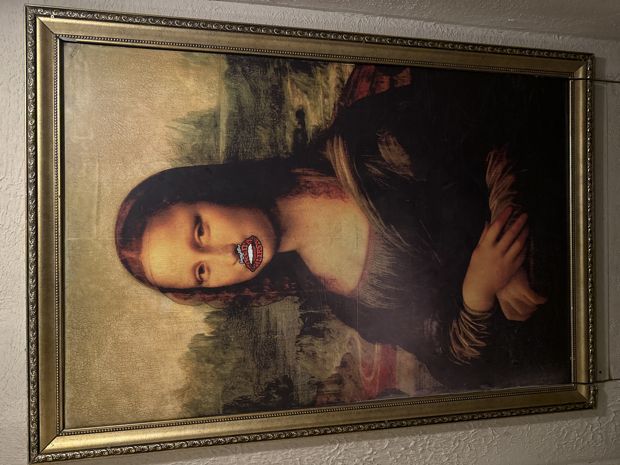 An altered portrait of the Mona Lisa, rotated 90 degrees counter-clockwise, is depicted in a medium shot. The artwork is framed in an ornate, golden frame with decorative patterns. The painting itself shows signs of craquelure, indicating age or an aged aesthetic.
The central subject, Mona Lisa, has her original mouth obscured by a superimposed, cartoonish image of open red lips with white teeth. A distinctive, shiny detail, resembling a diamond or "grill," is visible on the upper teeth within these superimposed lips. Inside the mouth, the text "Lounge Bar" is clearly readable. The background landscape and Mona Lisa's folded hands from the original painting are visible.
The framed painting is mounted on a textured, light-colored wall, possibly stucco or rough plaster, suggesting an indoor setting. A thin wire is visible near the upper right corner of the frame, indicating it is hung. The location context is Krasnoyarsk, Russia. No specific time of day or event is discernable beyond the display of this modified artwork.