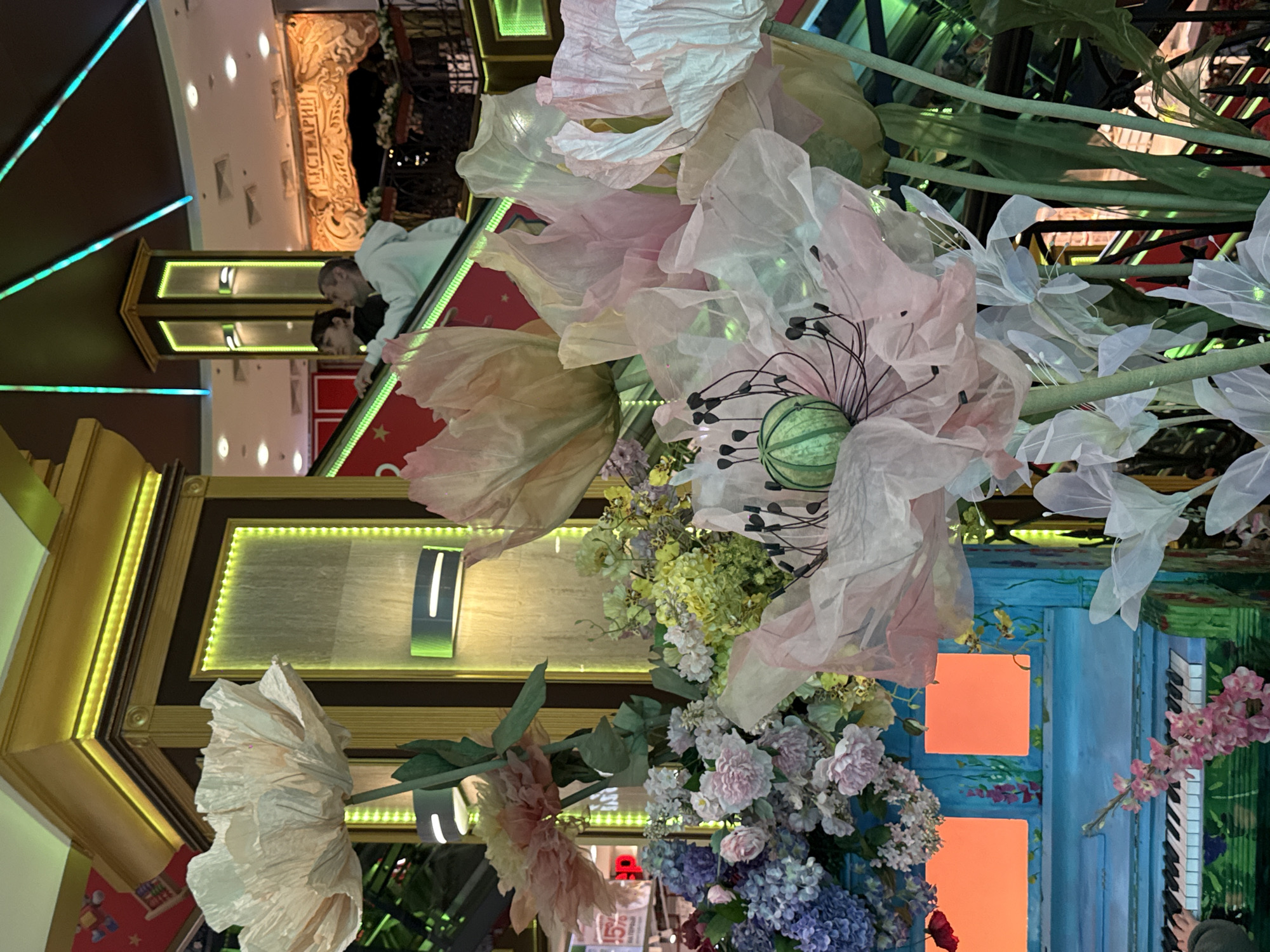 The photograph, taken in Moscow, Russia, presents an interior scene adorned with a whimsical arrangement of artificial flowers in shades of pink, white, and purple. These large-scale floral decorations dominate the foreground. Behind the floral display, architectural elements are visible, including a structure with golden accents and illuminated panels. People are situated on an escalator and can be seen through the flowers. A section of the scene resembles a painted backdrop, featuring a blue door with orange squares and a decorative piano keyboard.