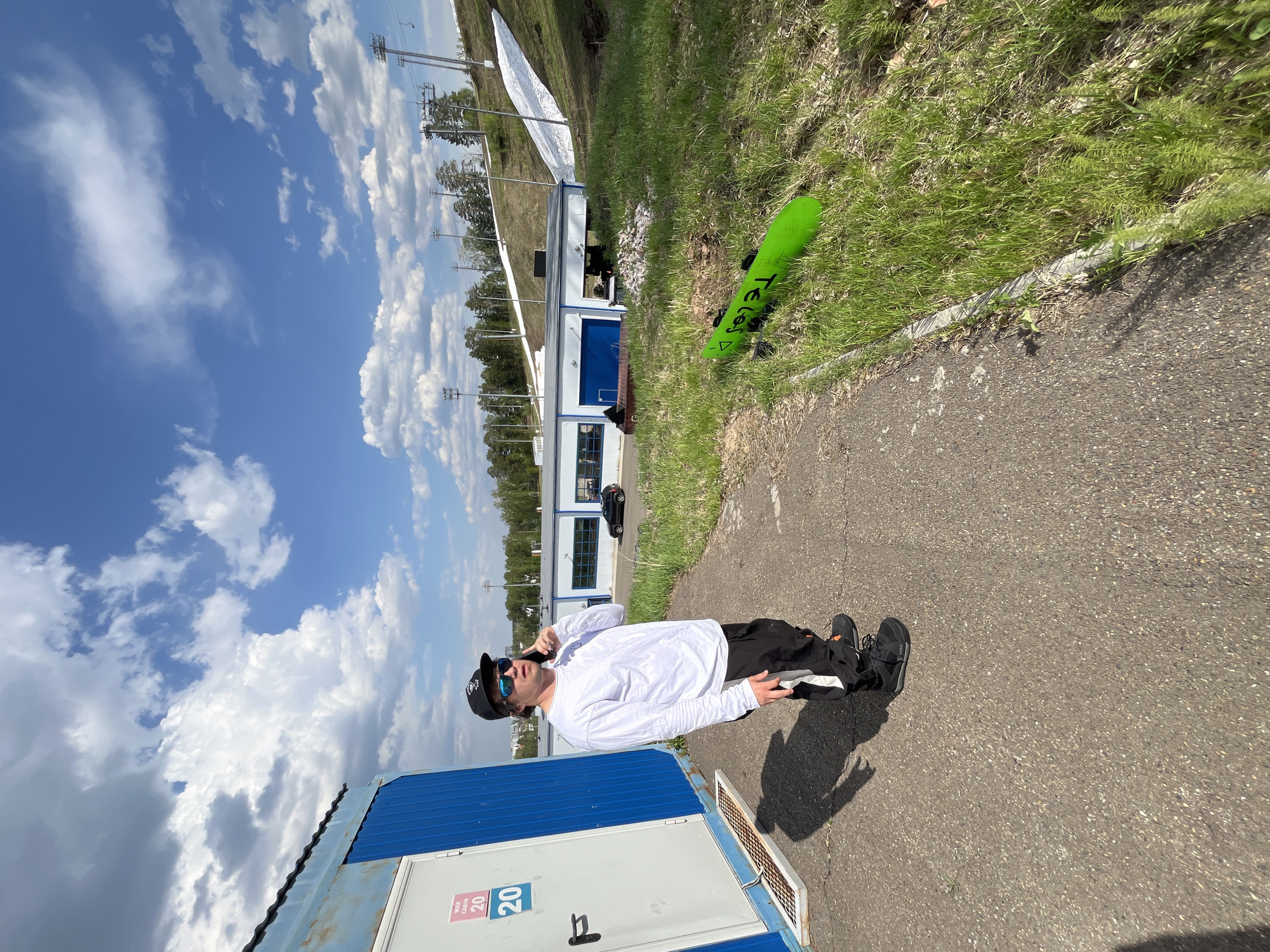 The image captures a man in Krasnoyarsk, Russia, standing on a paved surface near a blue and white modular building with the number "20" visible. He is wearing a white long-sleeved shirt, dark pants, black shoes, a cap, and sunglasses, and appears to be talking on a mobile phone.
In the background, there is a building, a snowy hill, and a green snowboard resting in the grass. The sky is blue with scattered clouds. The ground is asphalt.