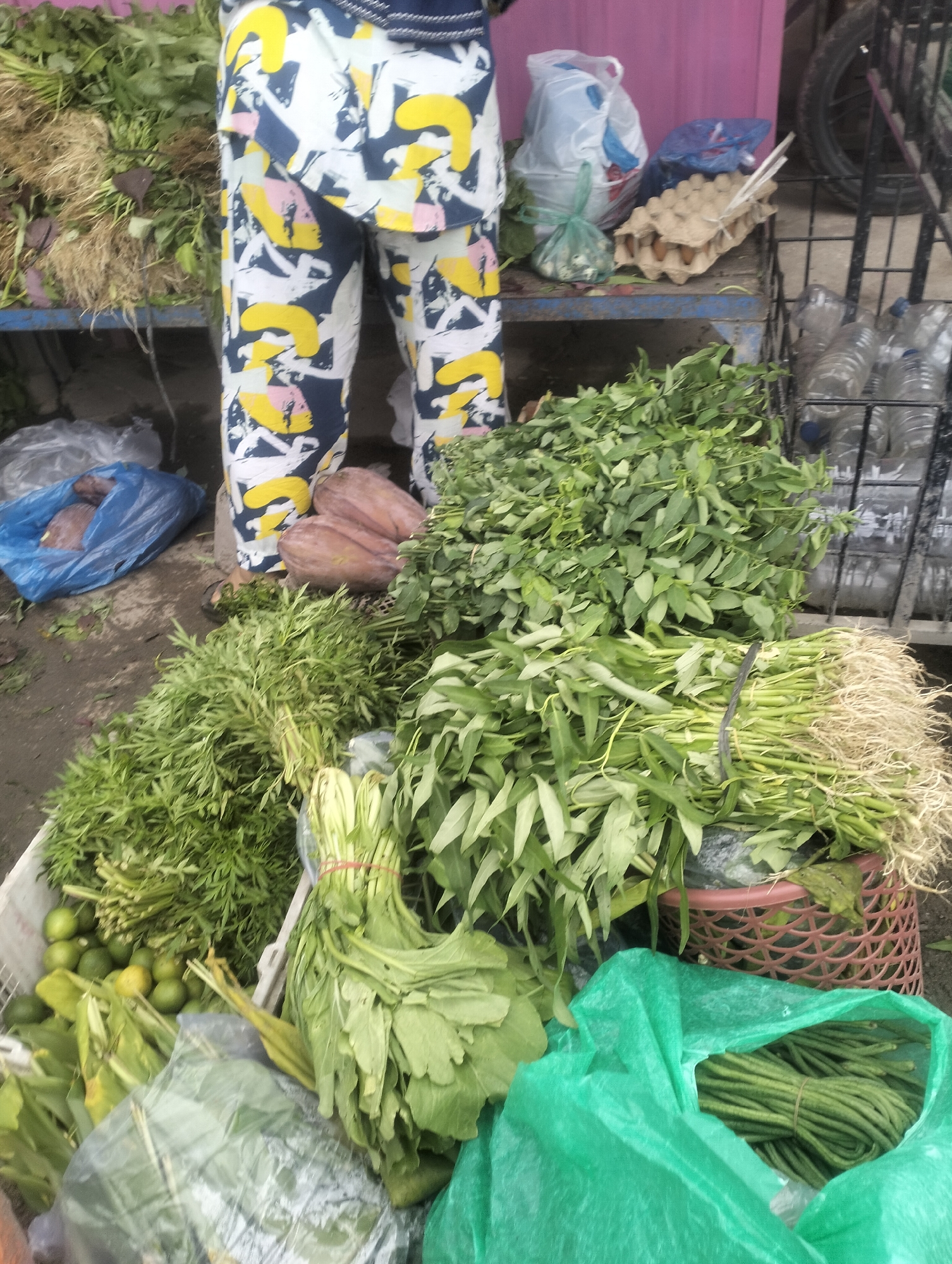 The image depicts a market stall in Dumai City, Indonesia, exhibiting a variety of fresh produce. In the foreground, a large bundle of green beans is visible inside a bright green plastic bag, secured with a rubber band. To the left, a white basket overflows with green vegetables, including leafy greens and small, round, green fruits resembling limes. Further back, several bunches of long, green vegetables with roots are arranged, possibly water spinach. A person's legs, clad in vibrantly patterned trousers with yellow, white, and navy blue designs, are partially visible. Behind the produce, an egg carton filled with eggs sits on a metal shelf, alongside plastic bags and bottles. The overall scene suggests a bustling market environment with a focus on fresh food items.