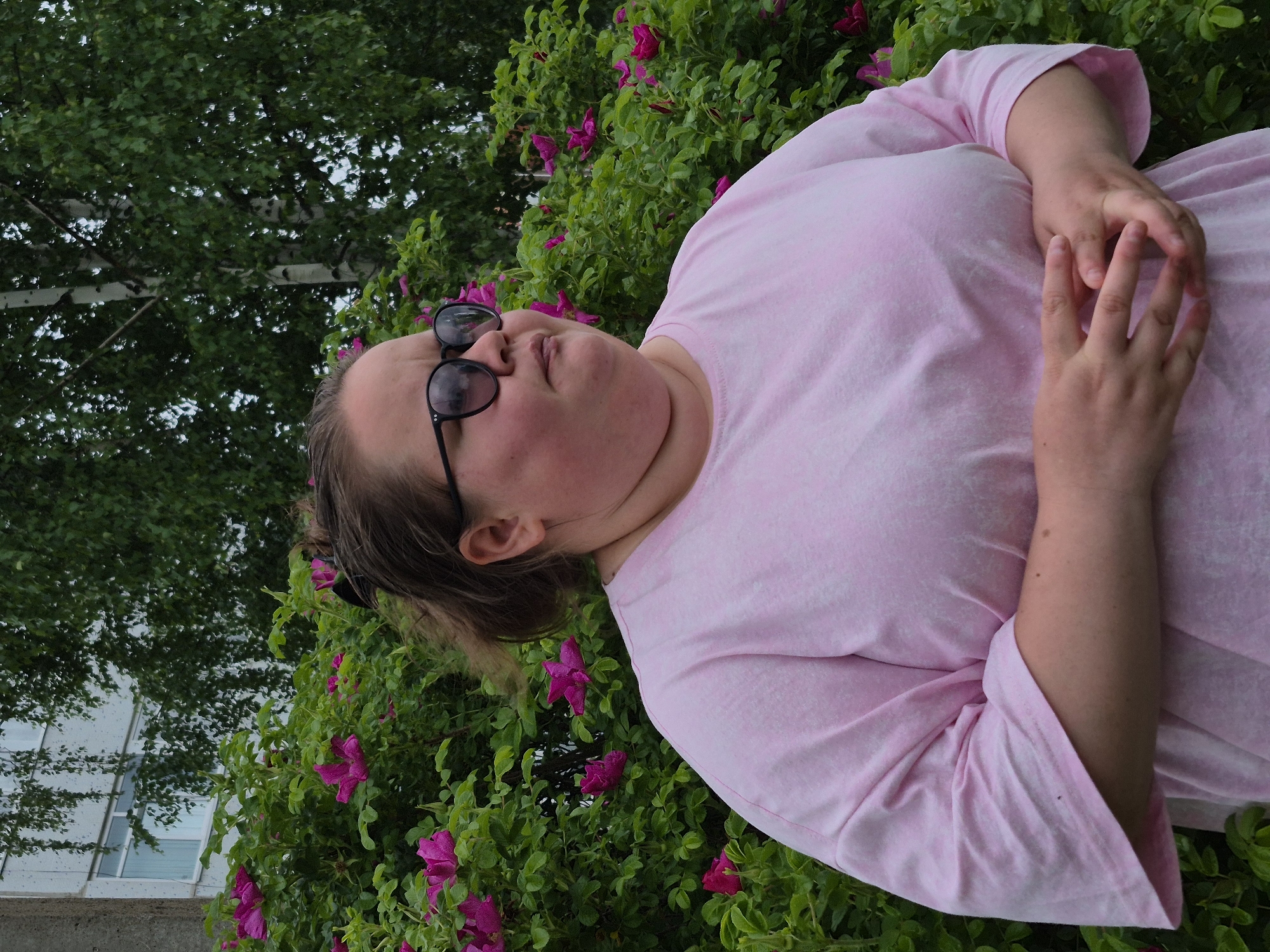 An individual, likely an adult female, is depicted from the chest up, posing outdoors. She wears a light pink, possibly tie-dye patterned, short-sleeved t-shirt. Black-framed sunglasses rest on her head. Her hands are gently clasped together in front of her chest, and her head is slightly tilted, with a neutral expression.
The subject is positioned in front of a vibrant green bush adorned with numerous magenta-pink flowers, resembling wild roses. Behind this flowering shrub, a large tree with green leaves and a light-colored trunk, characteristic of a birch tree, is visible in the upper left background. Further in the background, a modern building with multiple large glass windows is partially seen, hinting at an urban or semi-urban setting.
The scene takes place during daylight hours, with soft lighting suggesting an overcast sky or shaded area. The lush greenery and blooming flowers indicate spring or summer. The overall impression is a relaxed outdoor portrait set against a natural backdrop that borders an urban environment, consistent with a location such as Chaikovsky, Russia. There is no visible text in the image.
