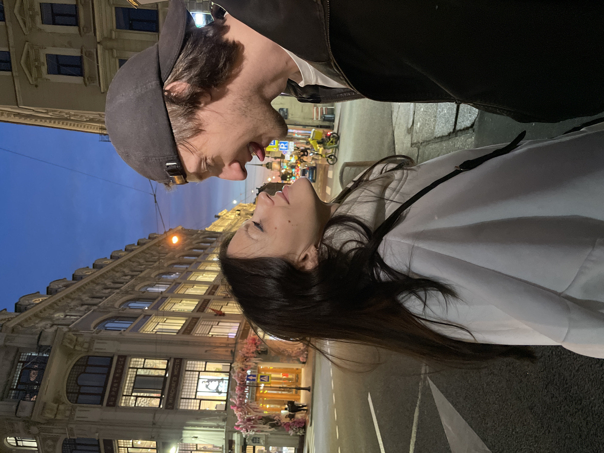 This is a medium shot, eye-level photograph taken at night in Saint Petersburg, Russia. The image features a couple in the foreground, facing each other and leaning in closely. The man on the right is wearing a dark jacket and a black baseball cap, and is sticking his tongue out towards the woman. The woman on the left has long dark hair and is wearing a white hooded jacket. Behind them, the street is illuminated by streetlights and the warm glow from the windows of a grand, ornate building, likely a department store, with signs visible in Russian. The sky is a deep twilight blue. A streetcar track is visible on the wet asphalt road, indicating an urban setting.